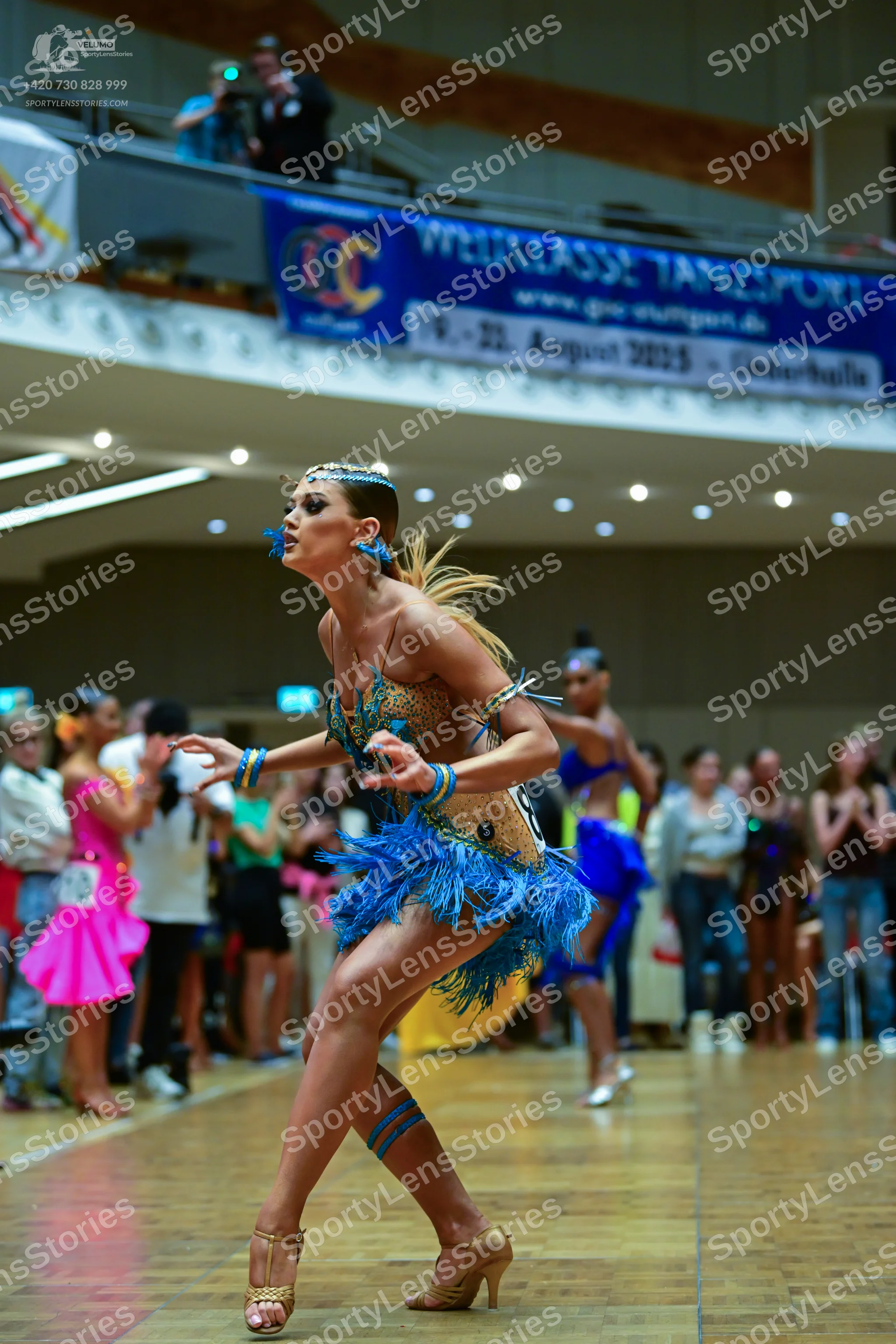Dancesport competitor: Foreground dancer: gold and blue Latin dress with sequins, blue fringe, and matc