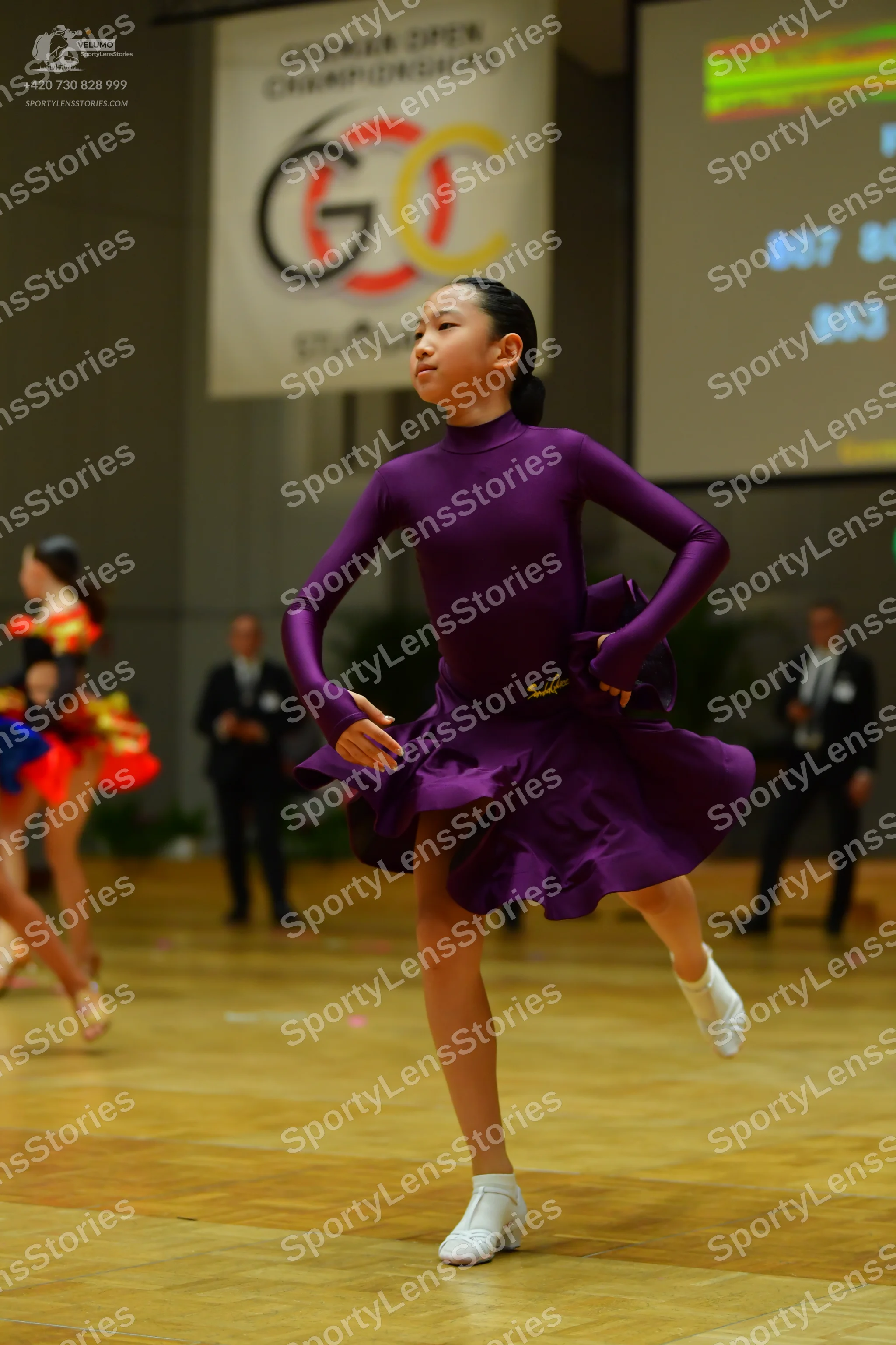 Dancesport competitor: Female dancer in foreground: purple long-sleeved latin dress with a high necklin
