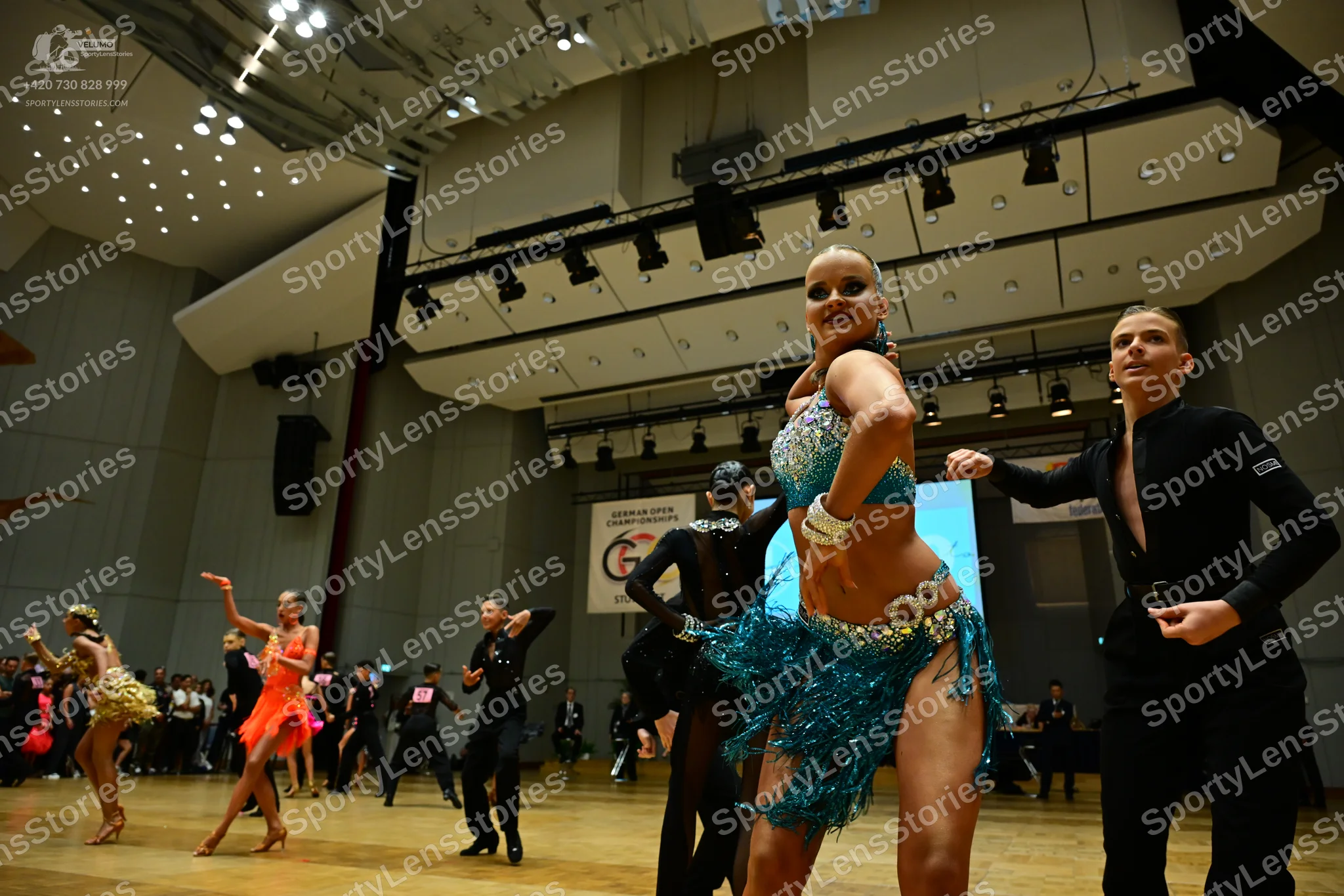 Dancesport competitor: Foreground female dancer: teal/turquoise sequined Latin dance costume with a seq