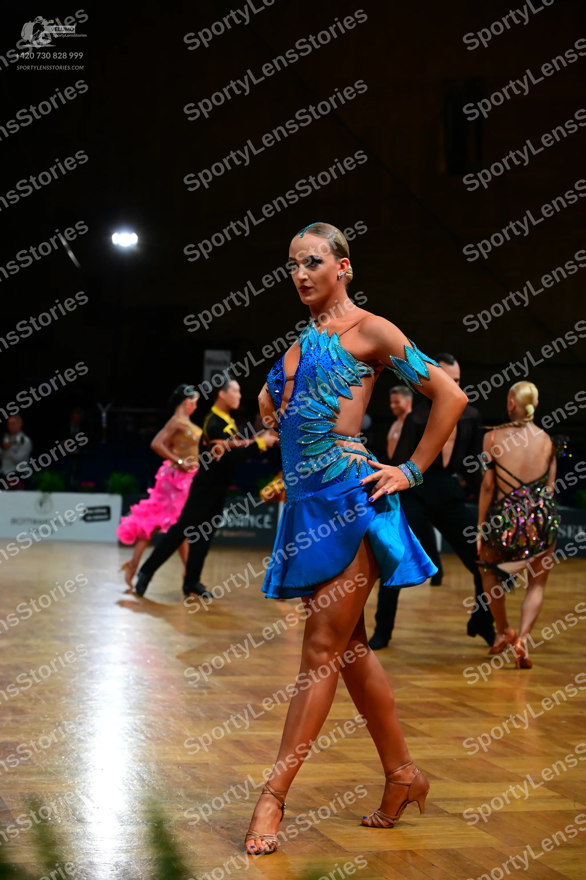 Dancesport competitor: Female dancer in foreground: blue latin dress with light blue/teal scale-like or