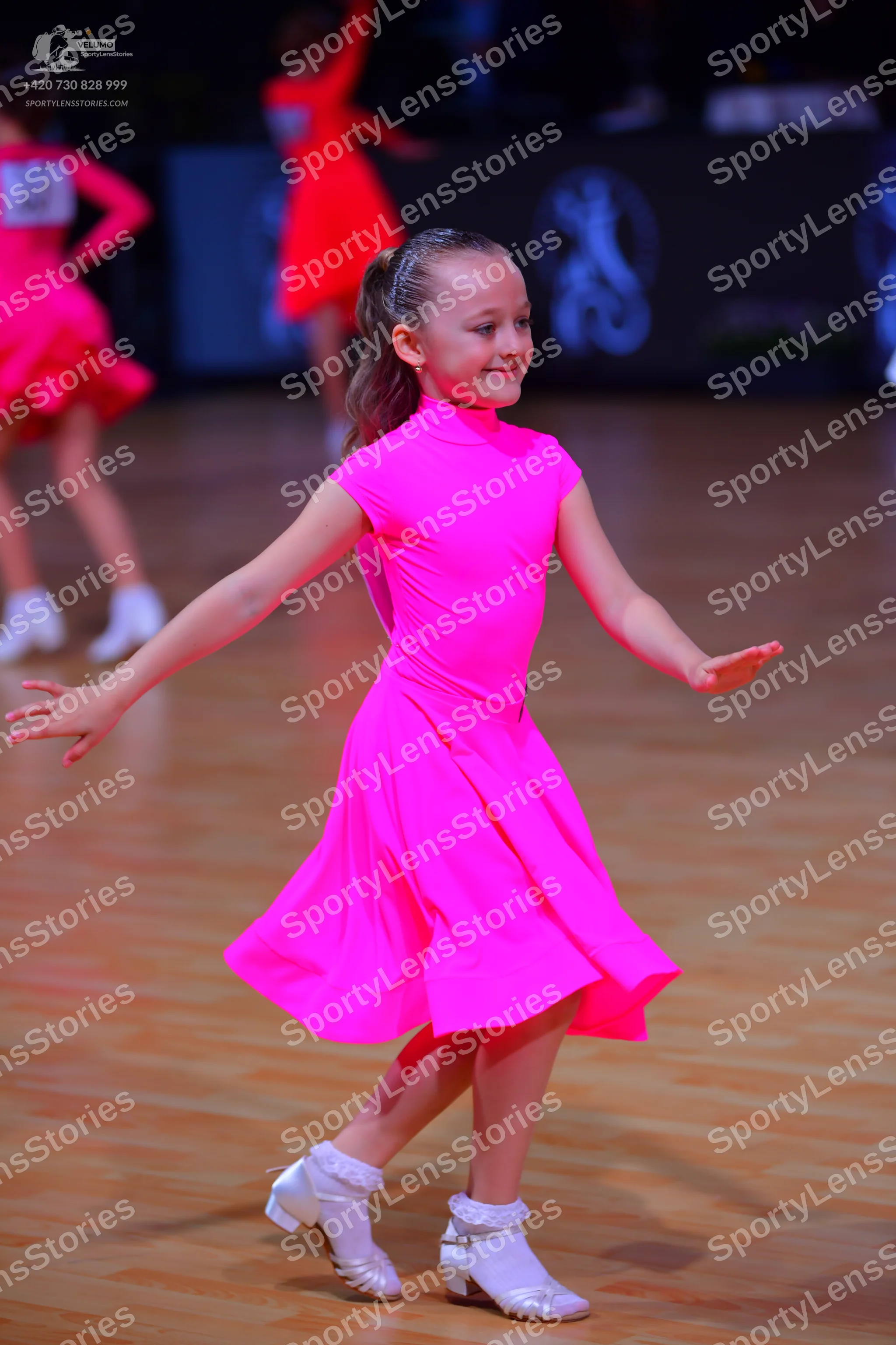 Dancesport competitor: Female dancer in foreground: bright pink short-sleeved latin dress with white da