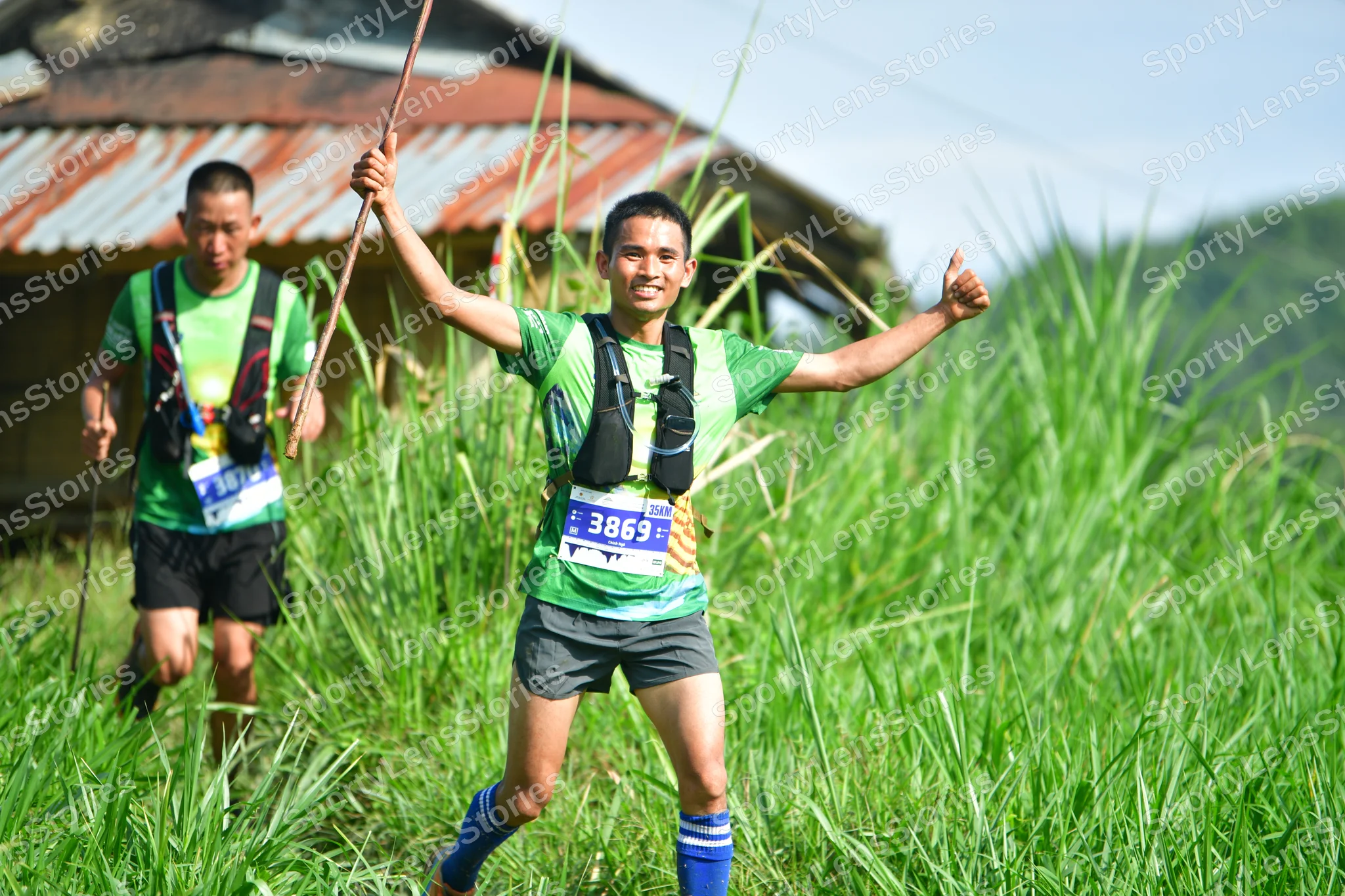 Marathon runner #3869, #3737: Runner 1: green patterned jersey, grey shorts, blue socks, black hydration vest