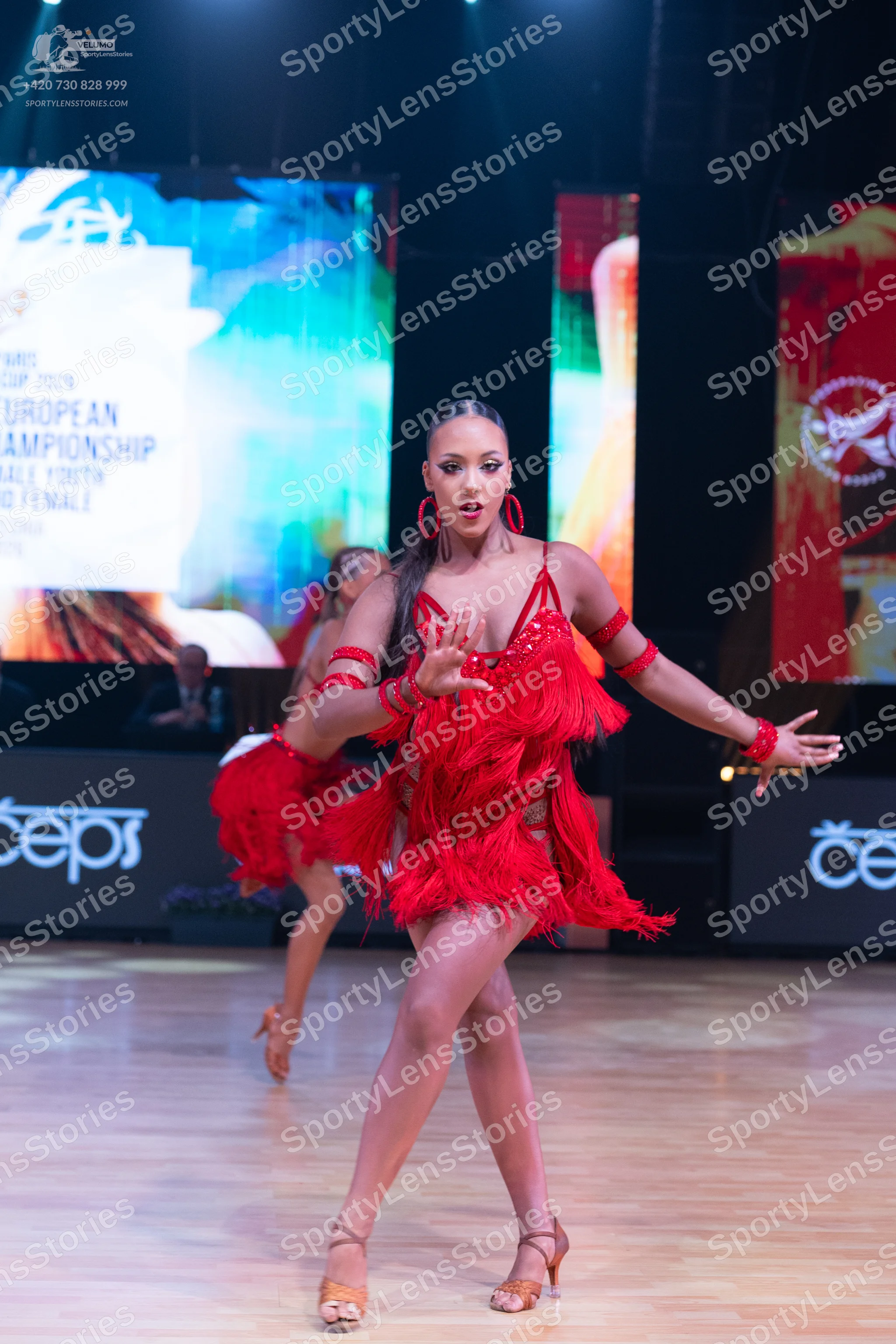 Dancesport competitor: Female dancer in foreground: red latin dress with fringe, red armbands, and red