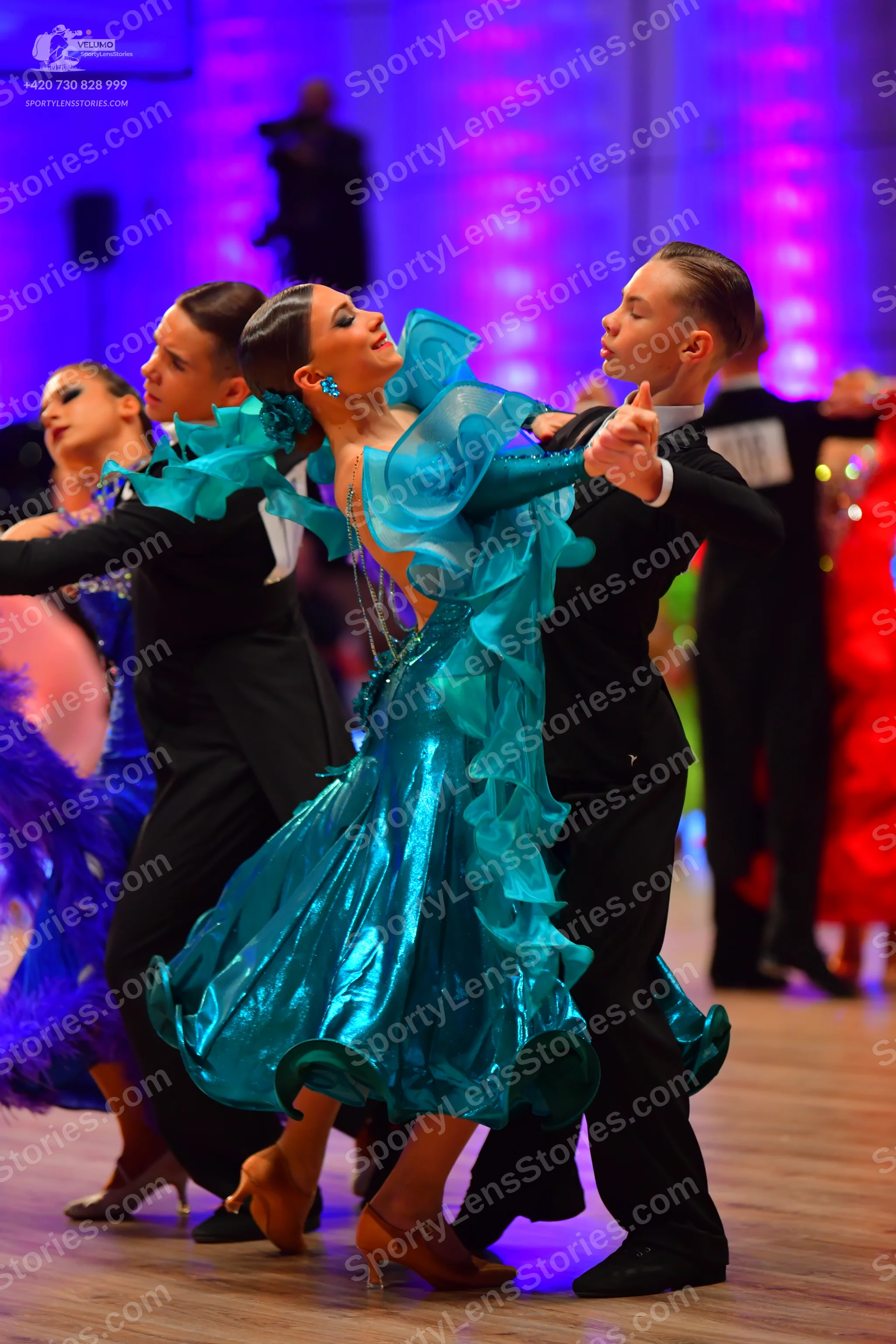 Dancesport competitor: Central female dancer: teal metallic Latin dress with large, flowing ruffled lay