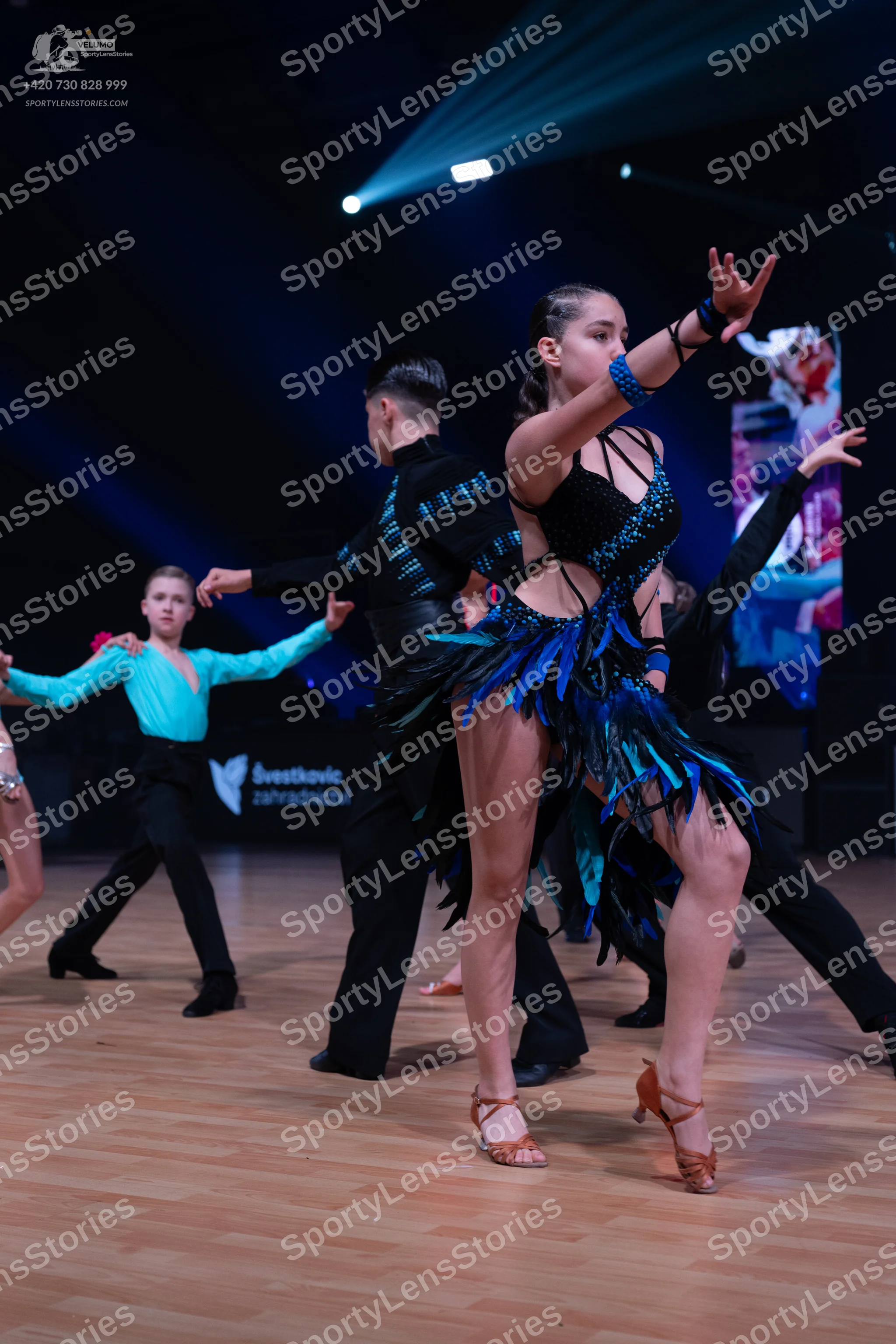 Dancesport competitor: Female dancer in foreground: black latin dress with blue and black feather-like
