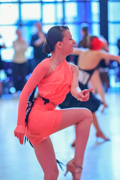 Dancesport competitor: Female dancer in foreground: peach-colored one-shoulder latin dress with black f