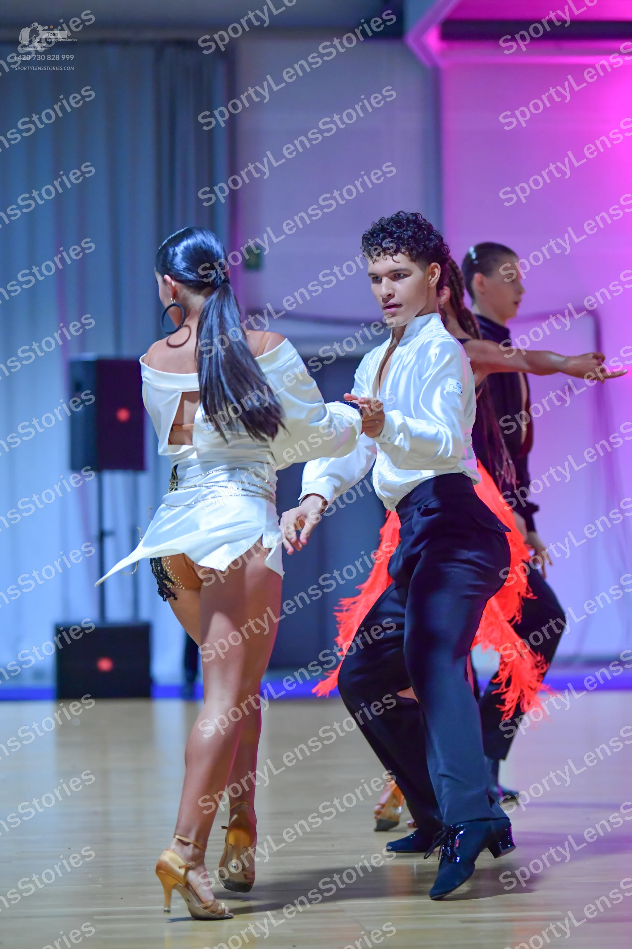 Dancesport competitor: Female dancer in foreground: white satin long-sleeved off-the-shoulder top and s