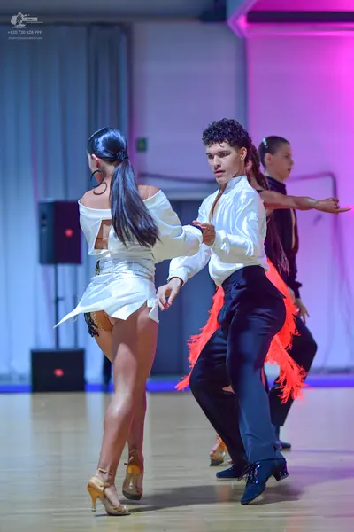 Dancesport competitor: Female dancer in foreground: white satin long-sleeved off-the-shoulder top and s