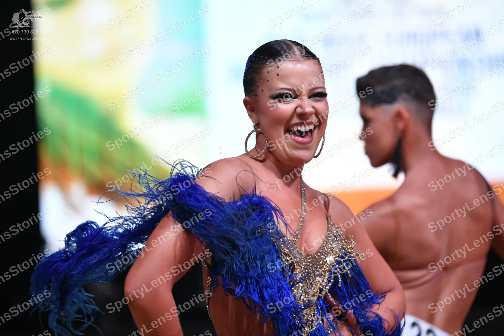Dancesport competitor: Female dancer: royal blue and gold latin dress with large blue feather accents o