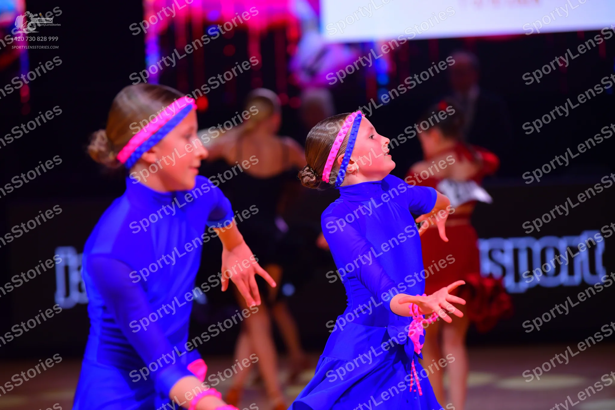 Dancesport competitor: Two dancers in matching blue long-sleeved dresses with pink headbands; one dance