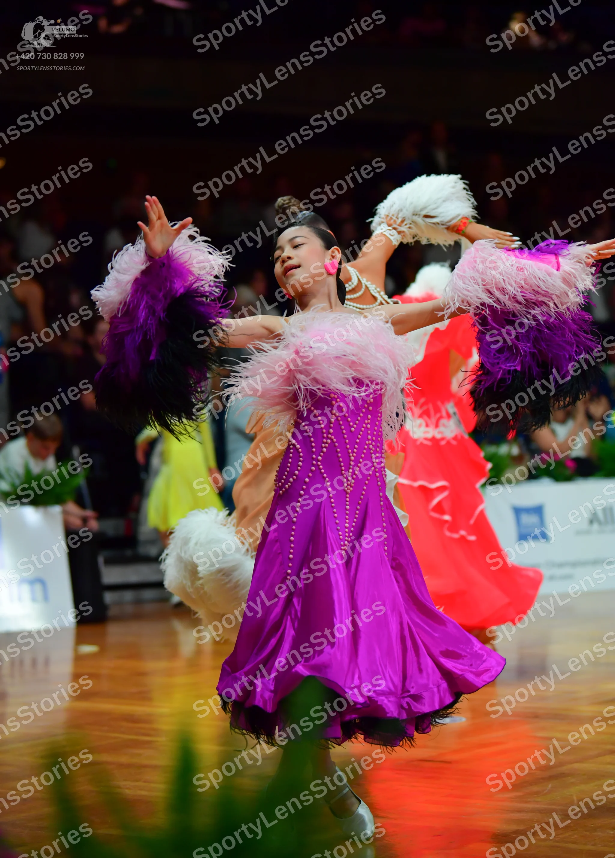 Dancesport competitor: Female dancer 1: Purple satin Latin dress featuring voluminous white, pink, and
