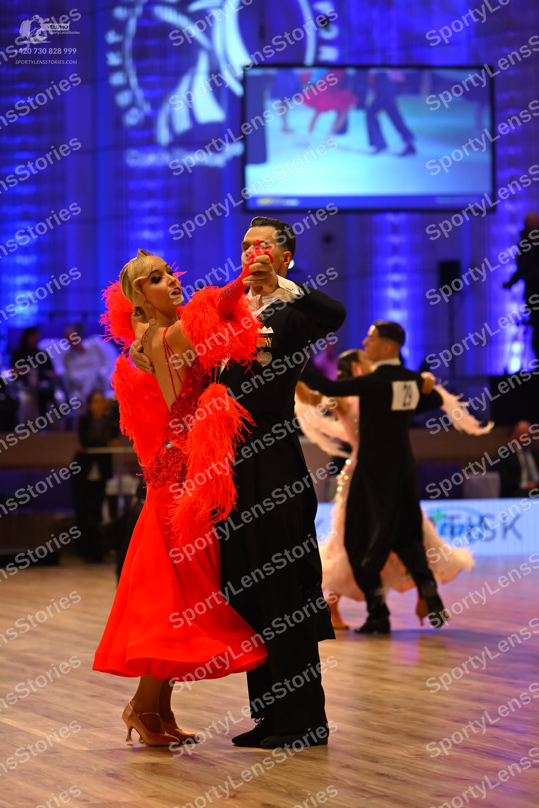 Dancesport competitor: Foreground female dancer: red Latin dress with large red feather/fringe accents