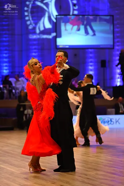 Dancesport competitor: Foreground female dancer: red Latin dress with large red feather/fringe accents 
