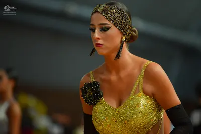Dancesport competitor: Female dancer: gold sequined latin dress with a black floral ornament on one sho
