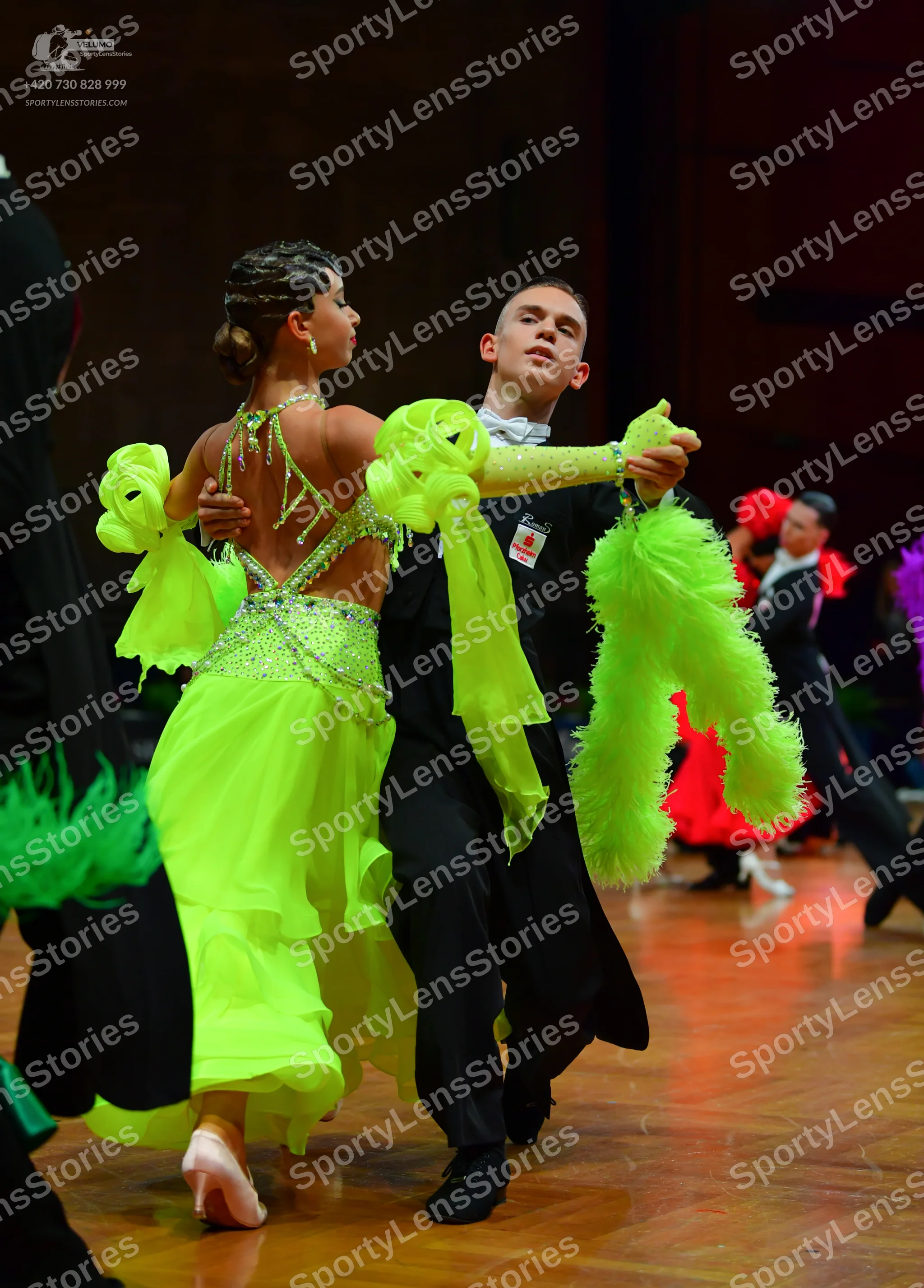 Dancesport competitor: Foreground female dancer: neon green Latin dress with sequins, ruffles, and a lo
