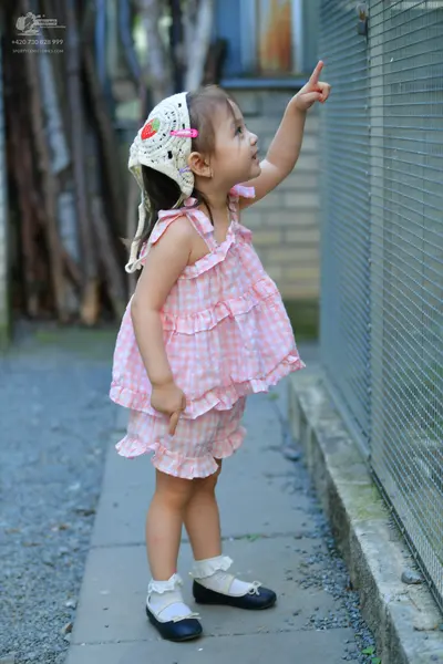 Dancesport competitor: The child is wearing a pink and white gingham sleeveless dress with ruffled laye
