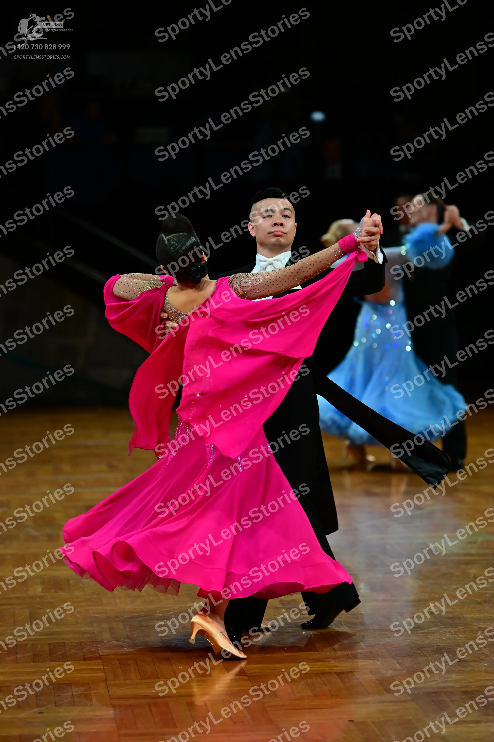 Dancesport competitor: Female dancer in foreground: bright pink flowing dress with a sheer, draped slee