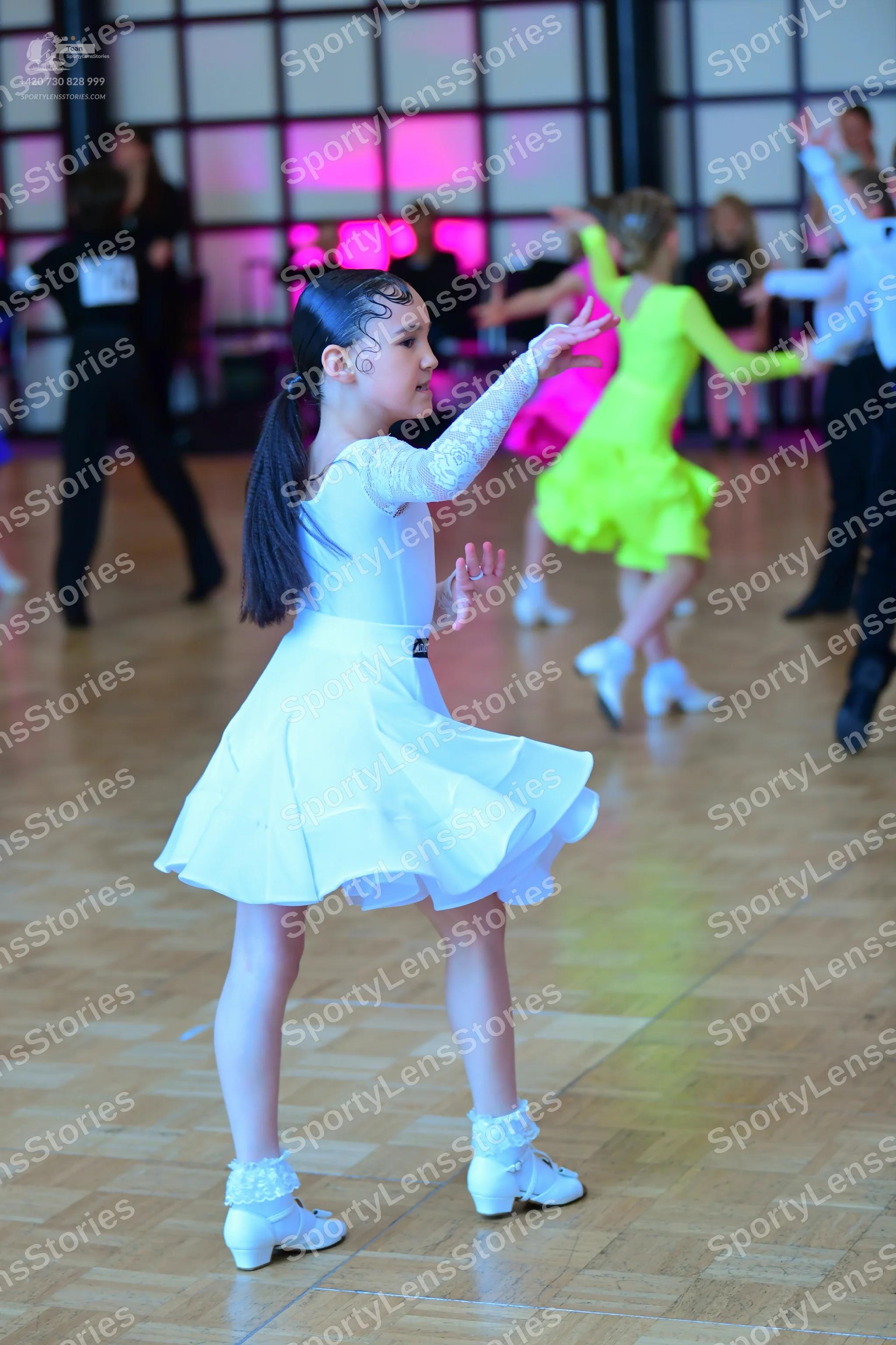 Dancesport competitor: Female dancer in foreground: white long-sleeved dress with lace sleeves and a fl