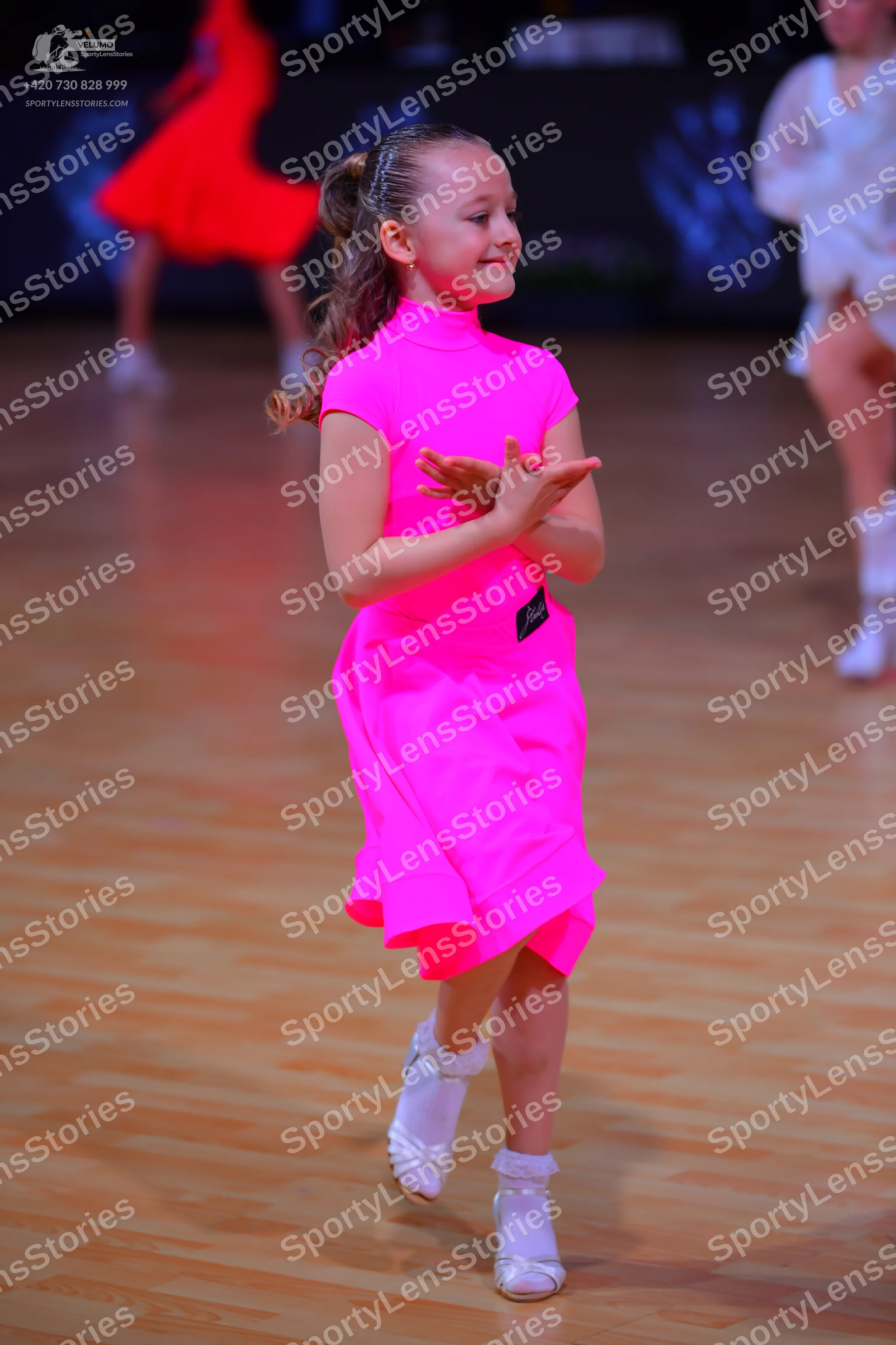 Dancesport competitor: Female dancer in foreground: bright neon pink short-sleeved dress with white dan