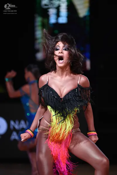 Dancesport competitor: Female dancer in foreground: black, yellow, orange, and pink latin dance costume