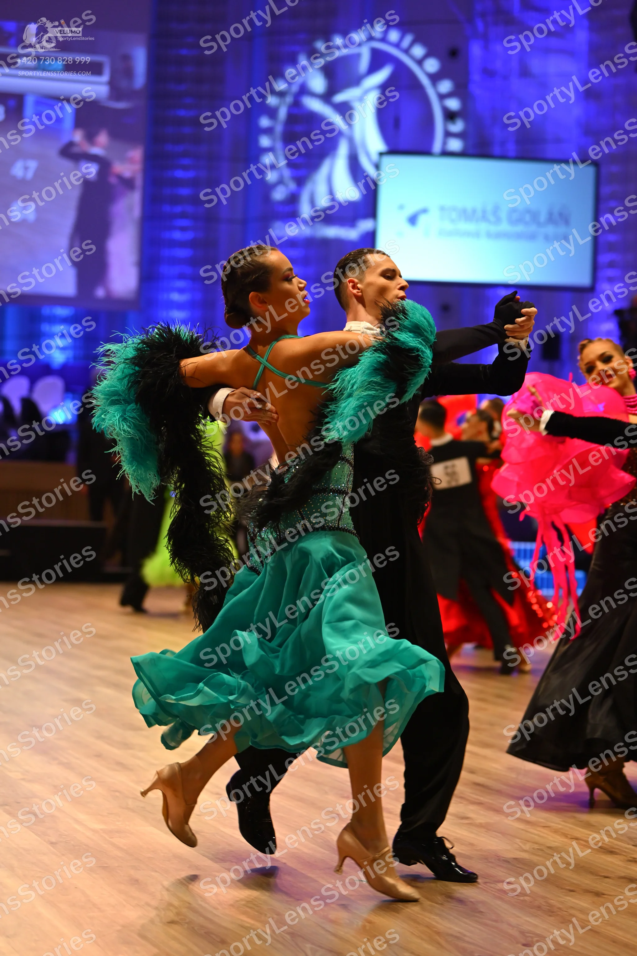 Dancesport competitor: Foreground female dancer: teal Latin dress with a sequined bodice, a flowing lay