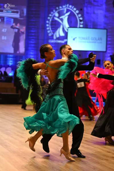 Dancesport competitor: Foreground female dancer: teal Latin dress with a sequined bodice, a flowing lay