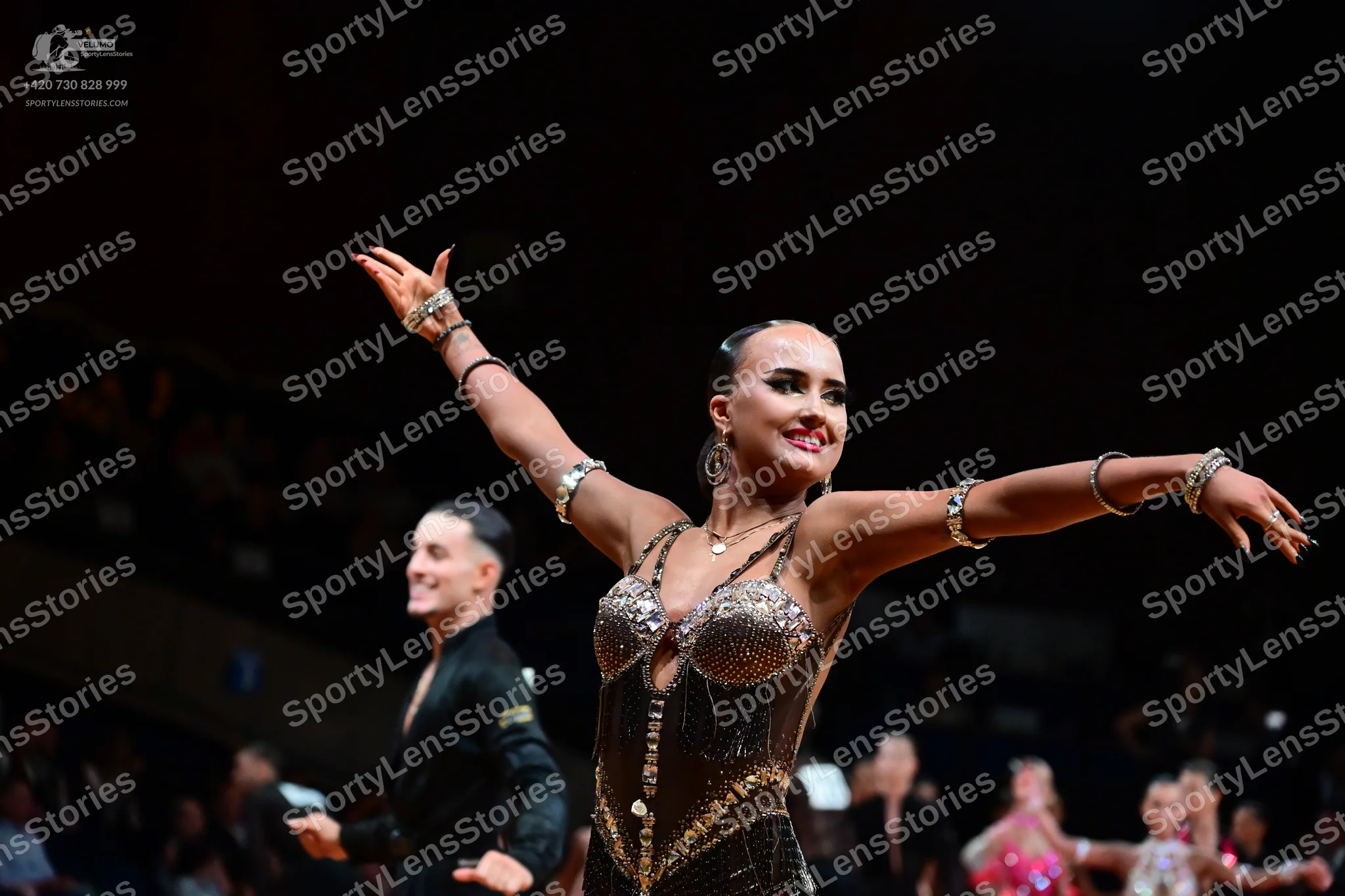 Dancesport competitor: Female dancer: dark, heavily embellished Latin dance bodysuit with bronze/black