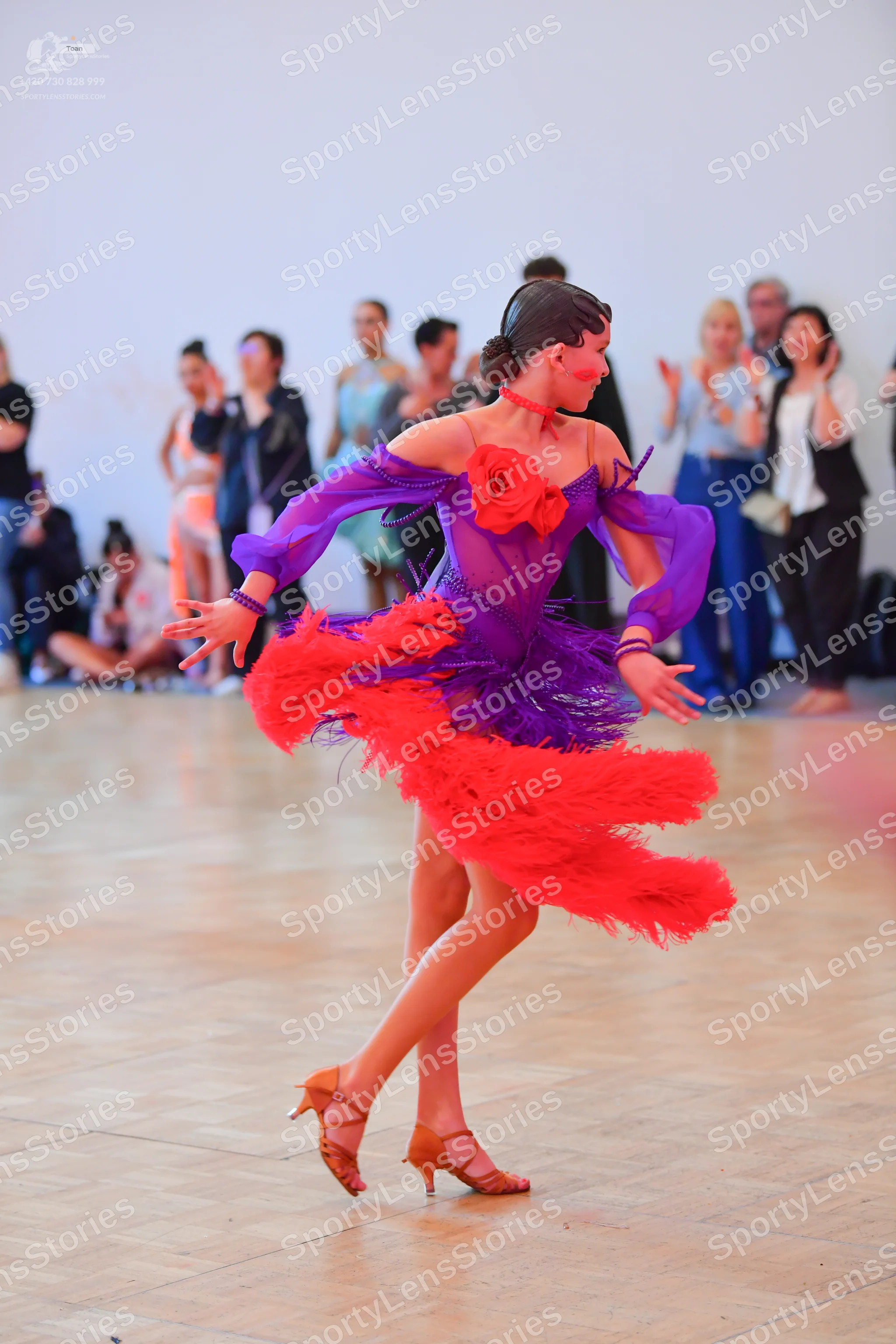 Dancesport competitor: Female dancer: purple latin dress with sheer sleeves, a large red rose on the bo