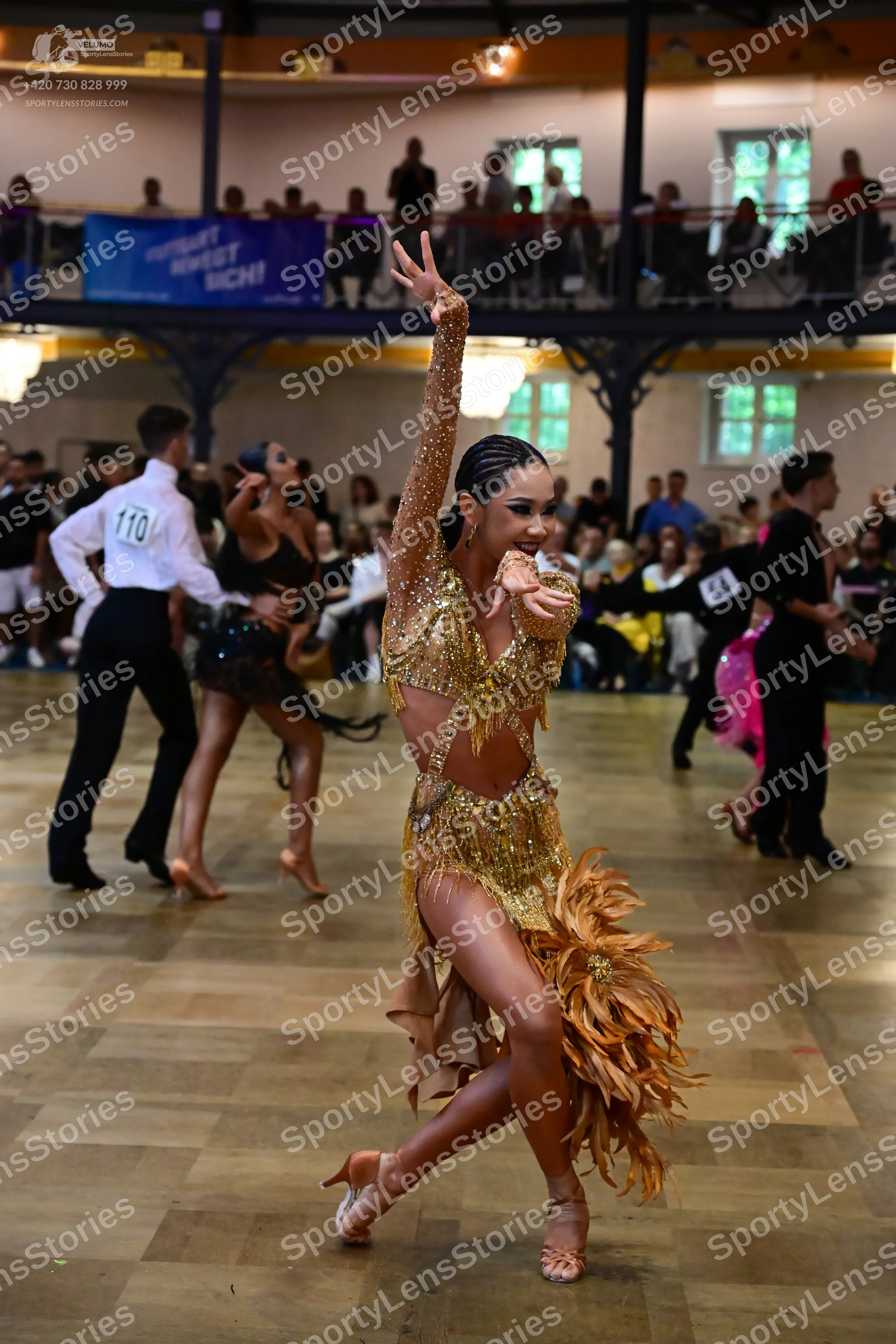 Dancesport competitor #110, #54: Female dancer in foreground: gold/bronze sequined latin dress with long, flowing