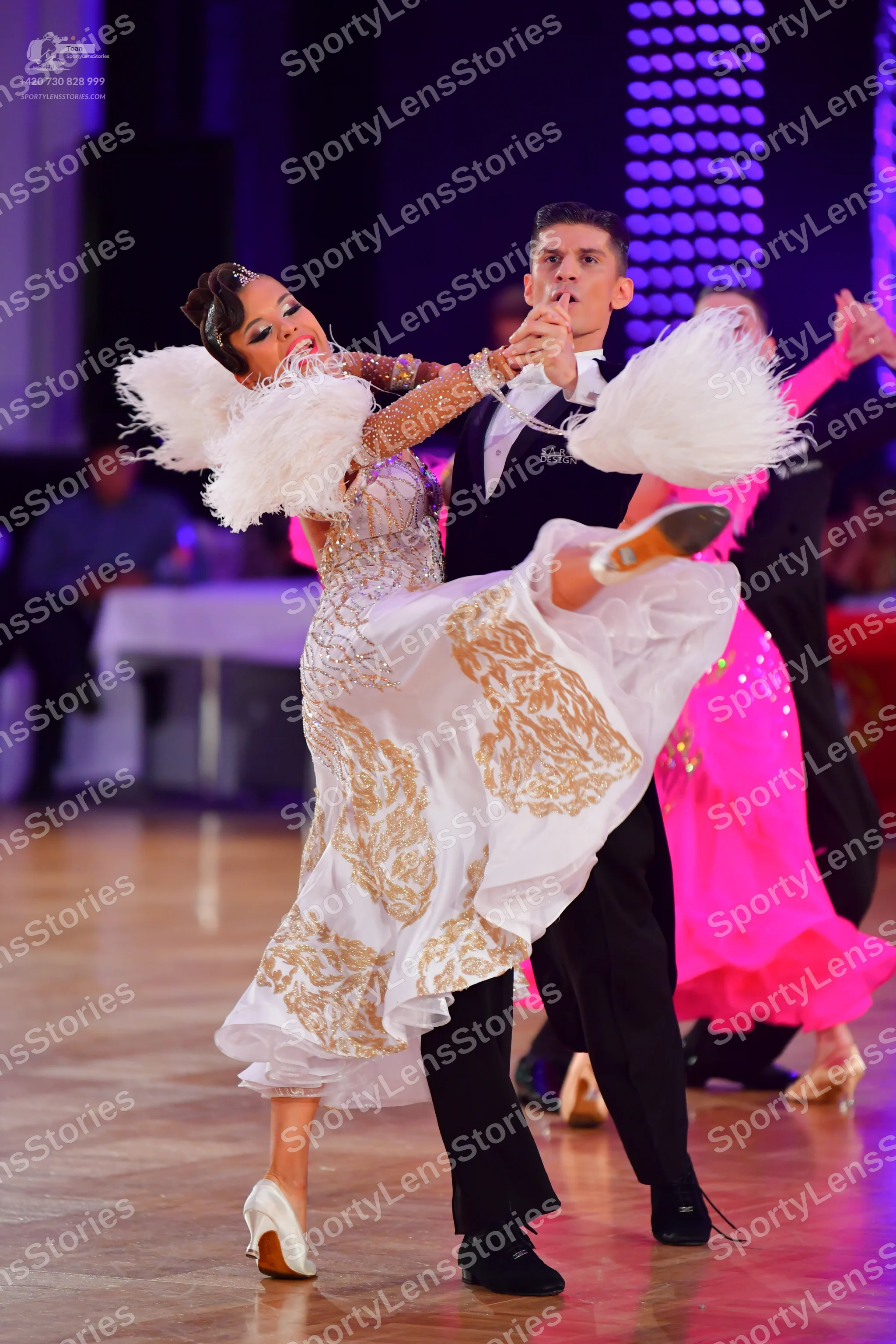 Dancesport competitor: Female dancer in foreground: white ballgown with gold sequined patterns and larg