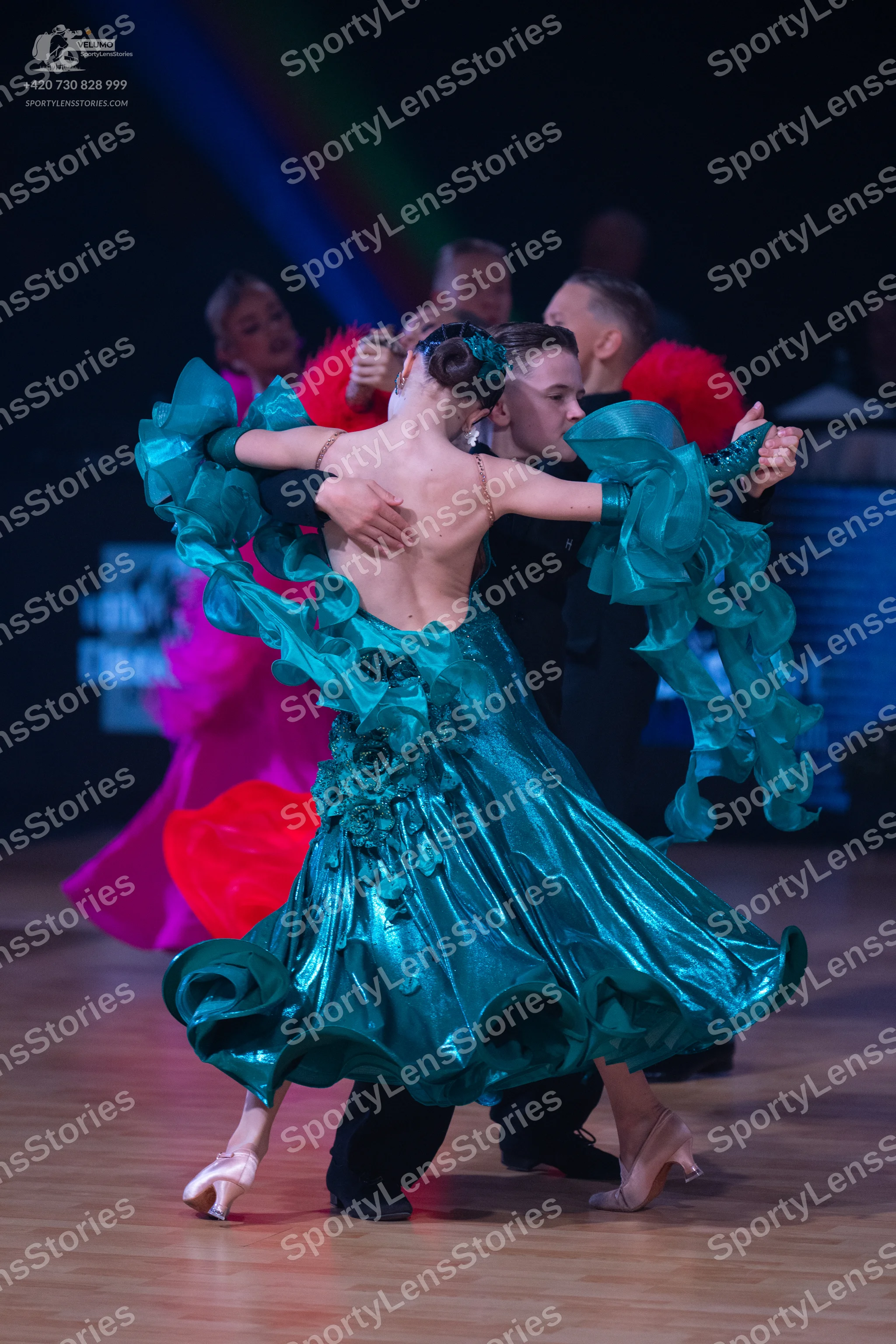 Dancesport competitor: Female dancer (foreground): teal ballroom dress with voluminous, ruffled layers 