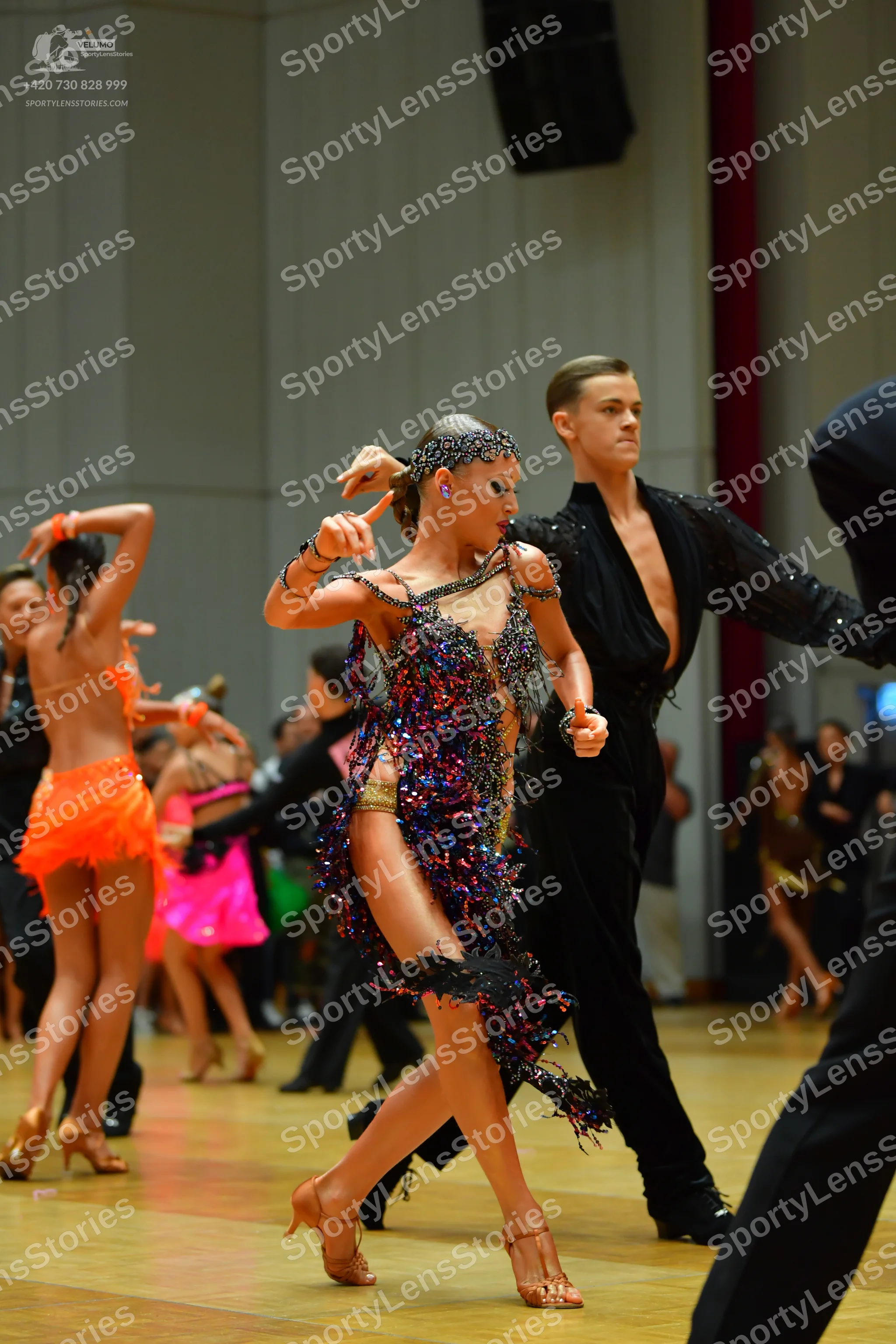 Dancesport competitor: Foreground female dancer: dark Latin dance dress heavily embellished with multi-