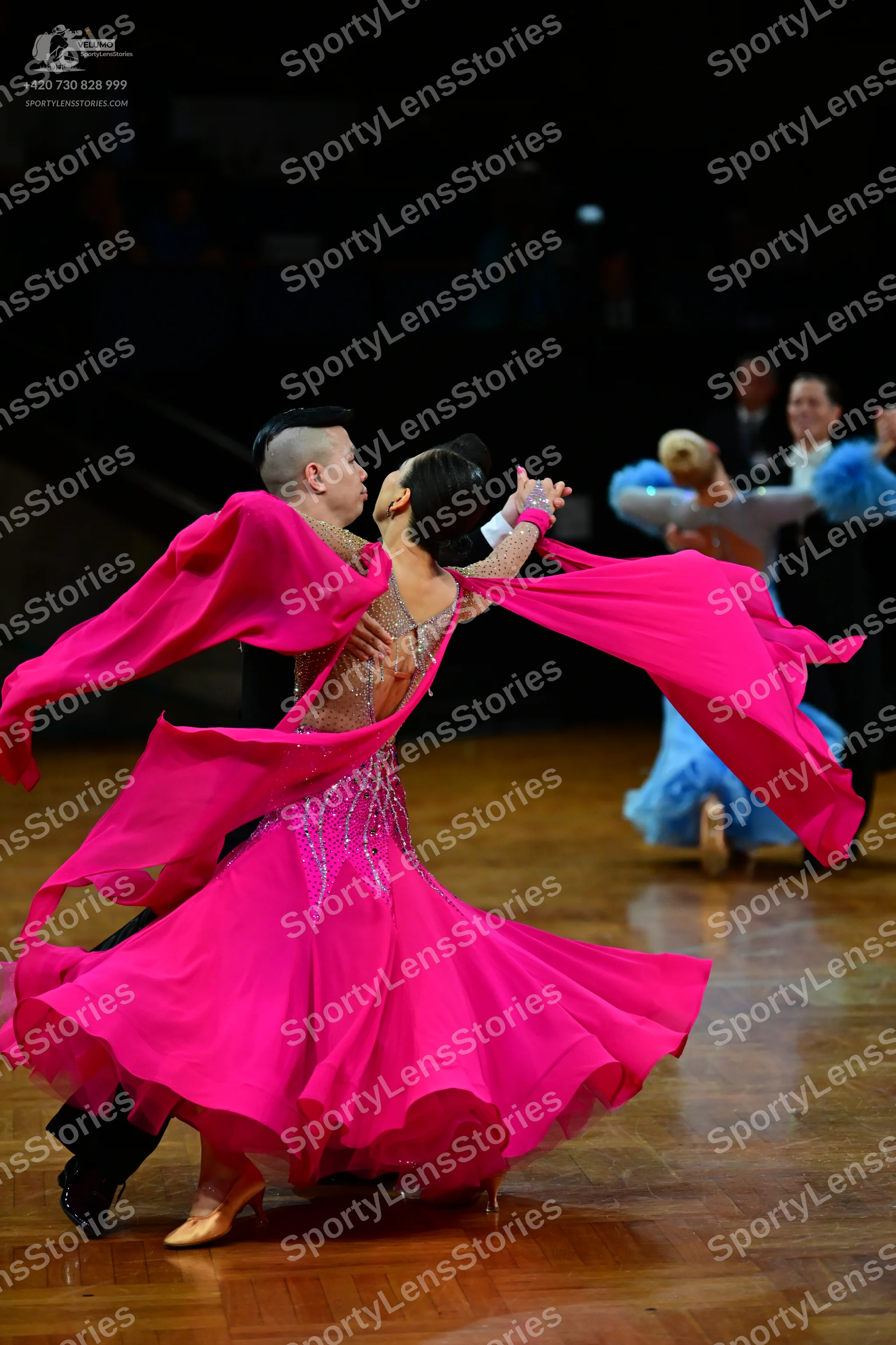 Dancesport competitor: Female dancer in foreground: bright pink ballroom dress with long, flowing sleev