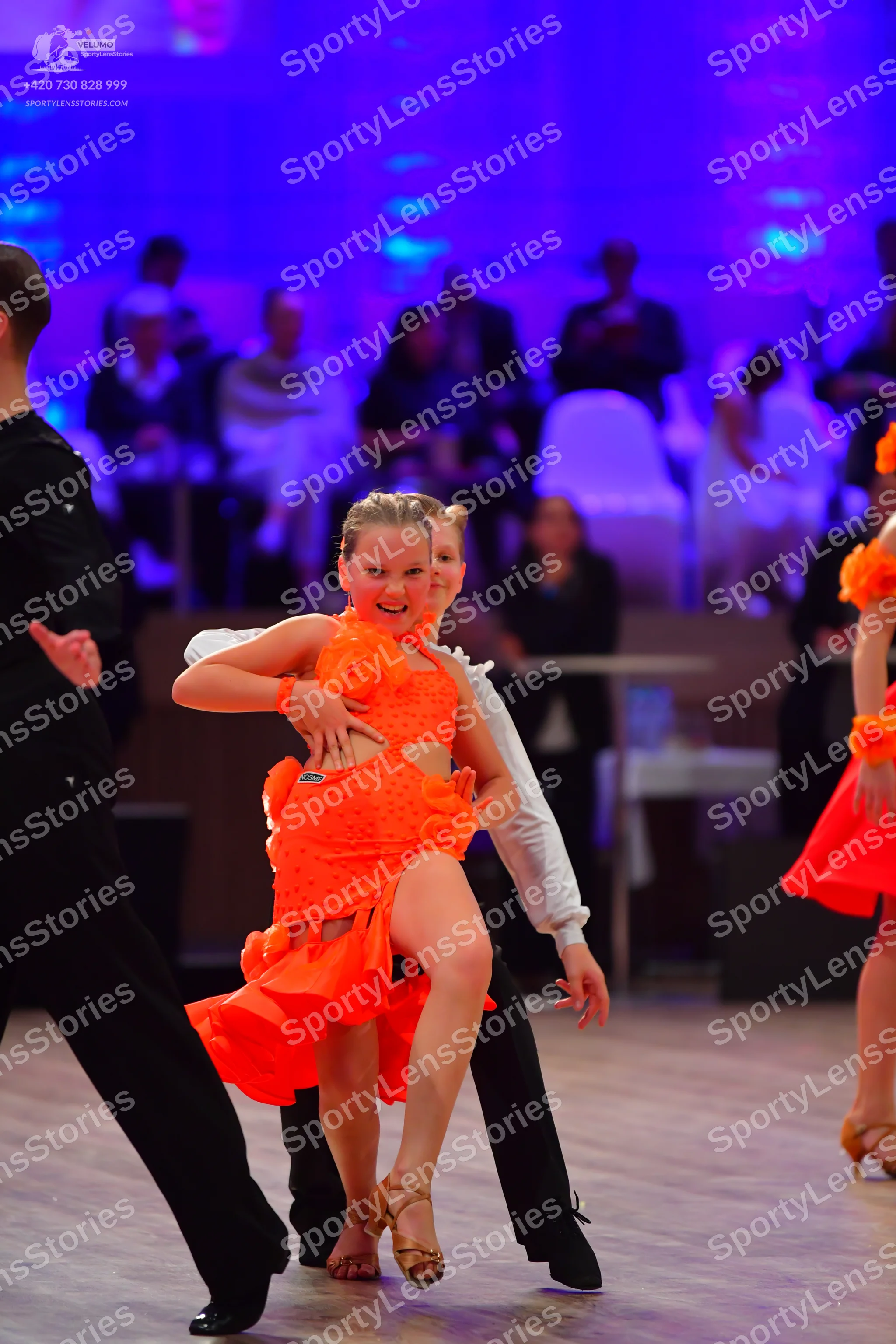 Dancesport competitor: Female dancer in foreground: orange Latin dress with ruffles and side cutouts