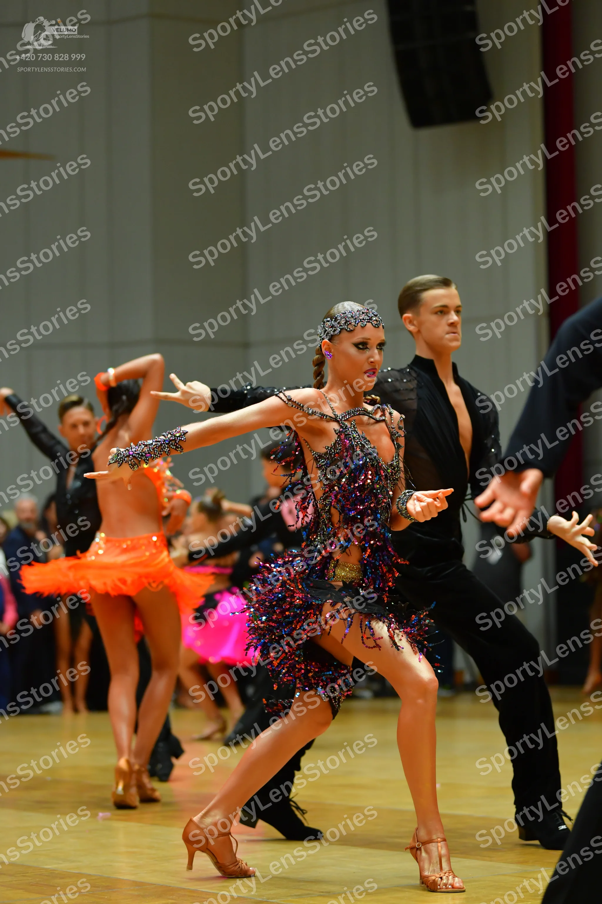 Dancesport competitor: Foreground female dancer: dark-colored (black, purple, and blue) Latin dance dre