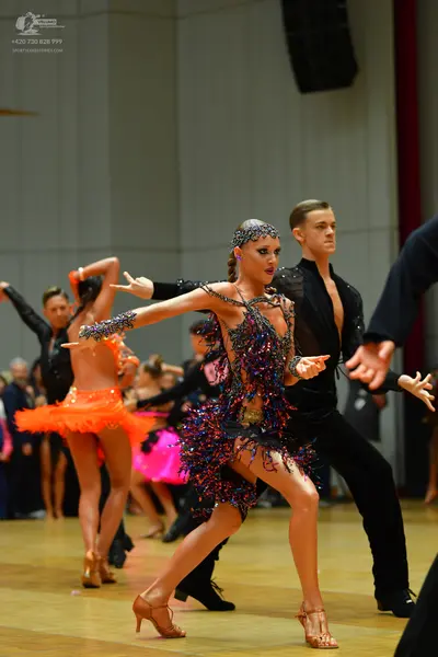 Dancesport competitor: Foreground female dancer: dark-colored (black, purple, and blue) Latin dance dre