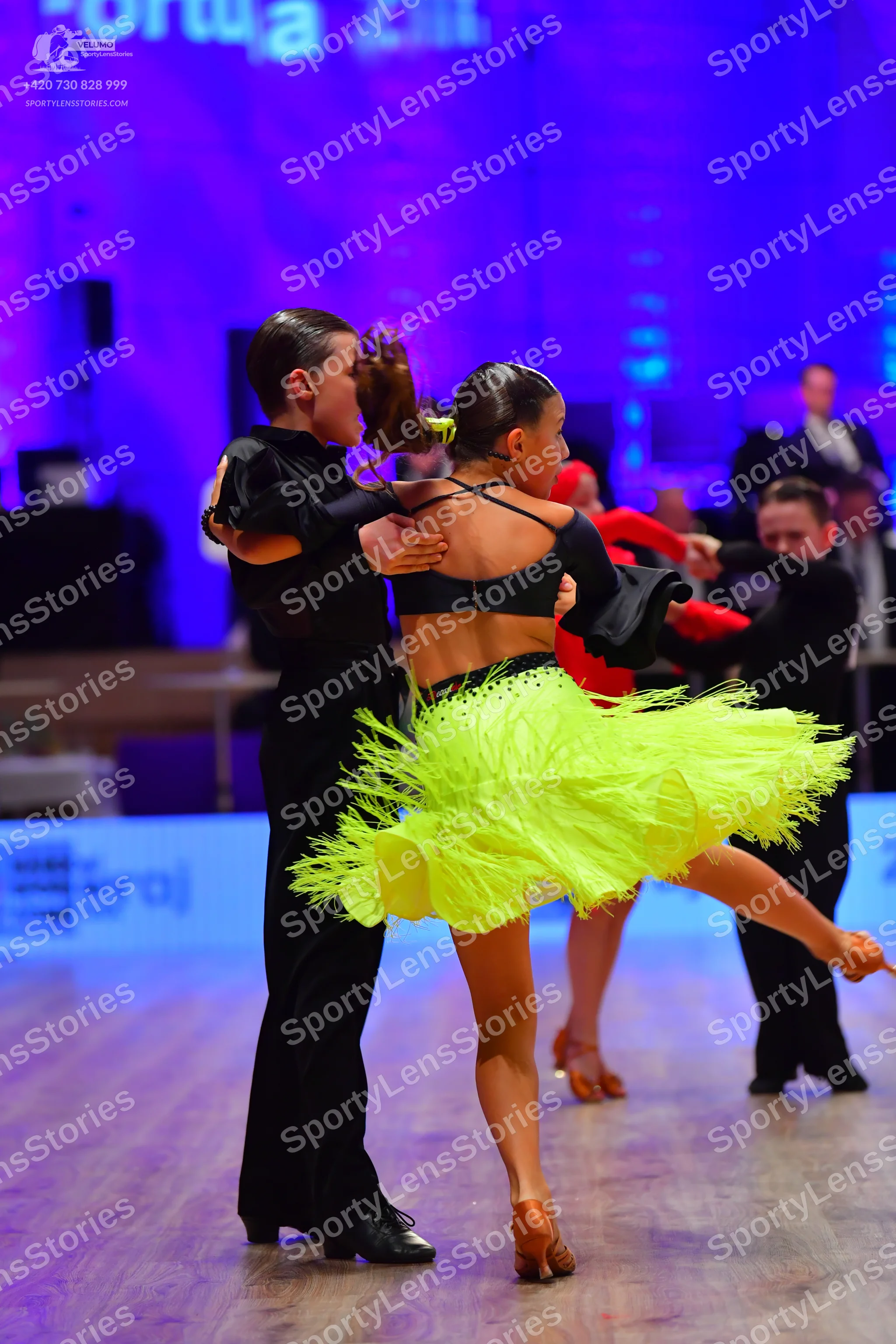 Dancesport competitor: Female dancer in foreground: black crop top and bright yellow fringe skirt