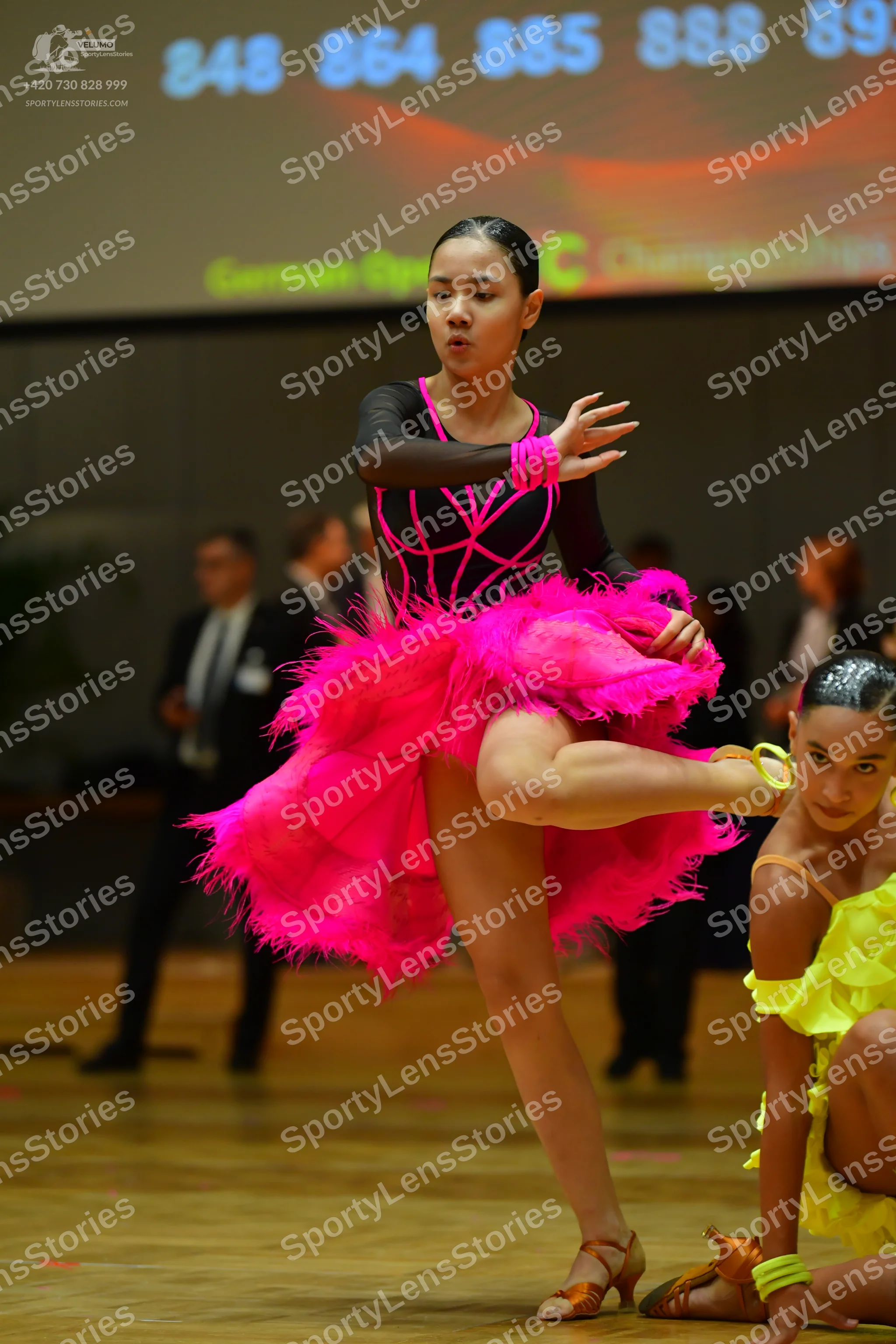 Dancesport competitor #848, #864: Female dancer 1: black long-sleeve top with pink trim and a bright pink feathere