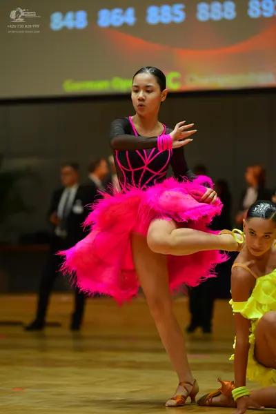 Dancesport competitor #848, #864: Female dancer 1: black long-sleeve top with pink trim and a bright pink feathere