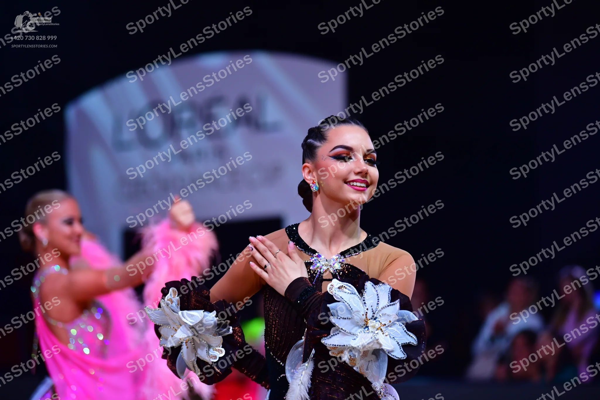 Dancesport competitor: Female dancer in foreground: black latin dance dress featuring large white fabri