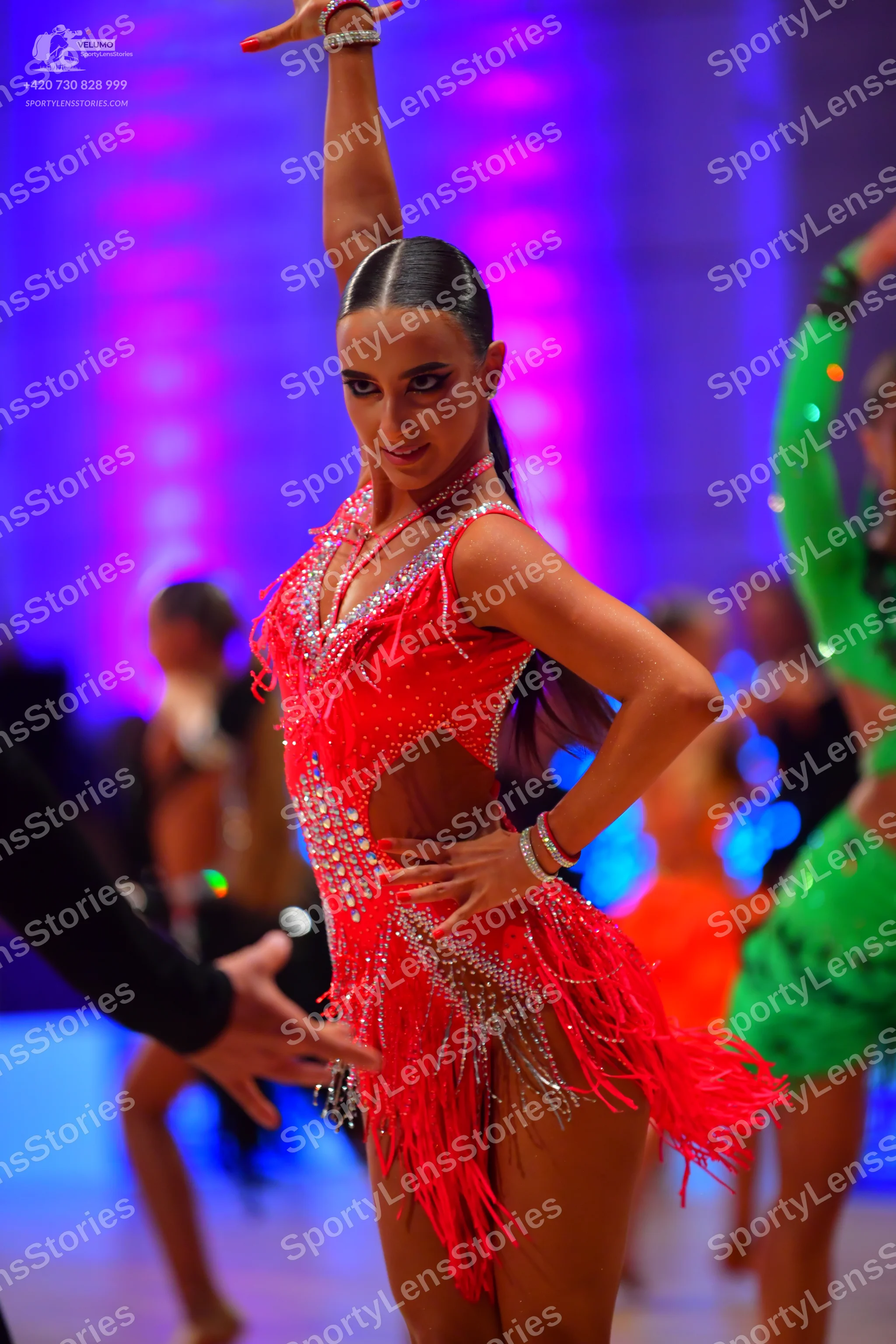 Dancesport competitor: Female dancer in foreground: red Latin dress with sequins, cutouts, and long fri