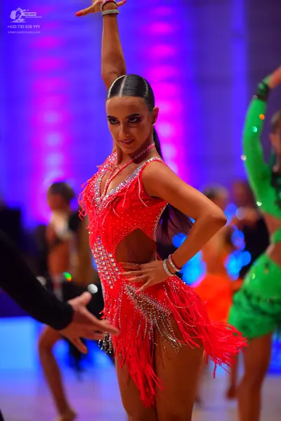 Dancesport competitor: Female dancer in foreground: red Latin dress with sequins, cutouts, and long fri