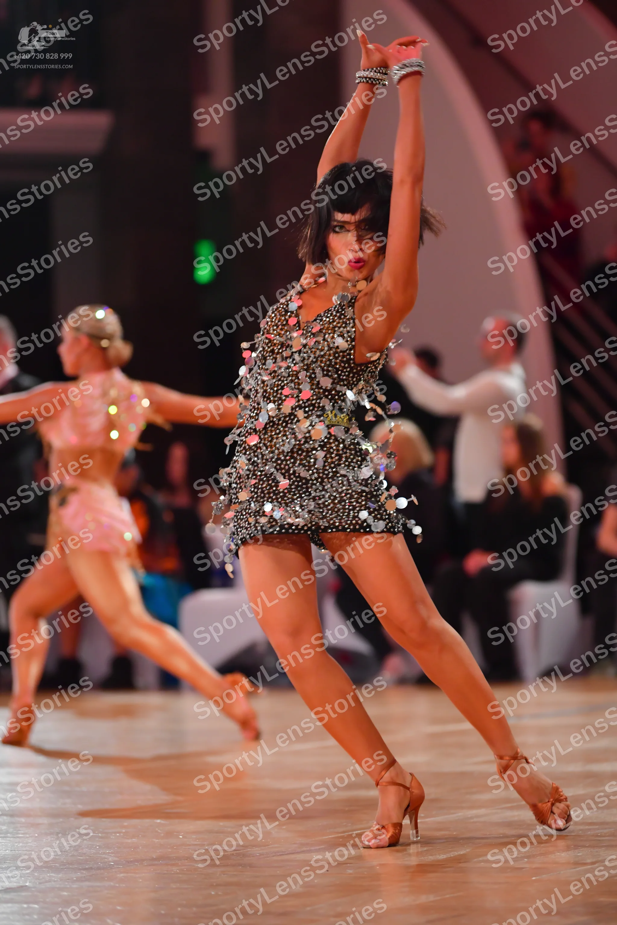 Dancesport competitor: Female dancer in foreground: dark-colored short Latin dress covered in multi-col