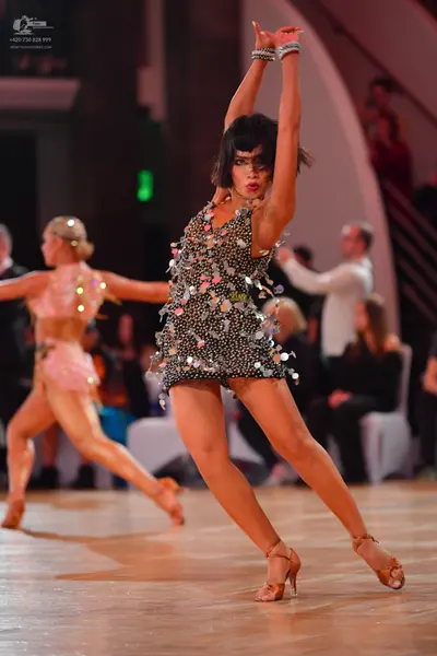 Dancesport competitor: Female dancer in foreground: dark-colored short Latin dress covered in multi-col