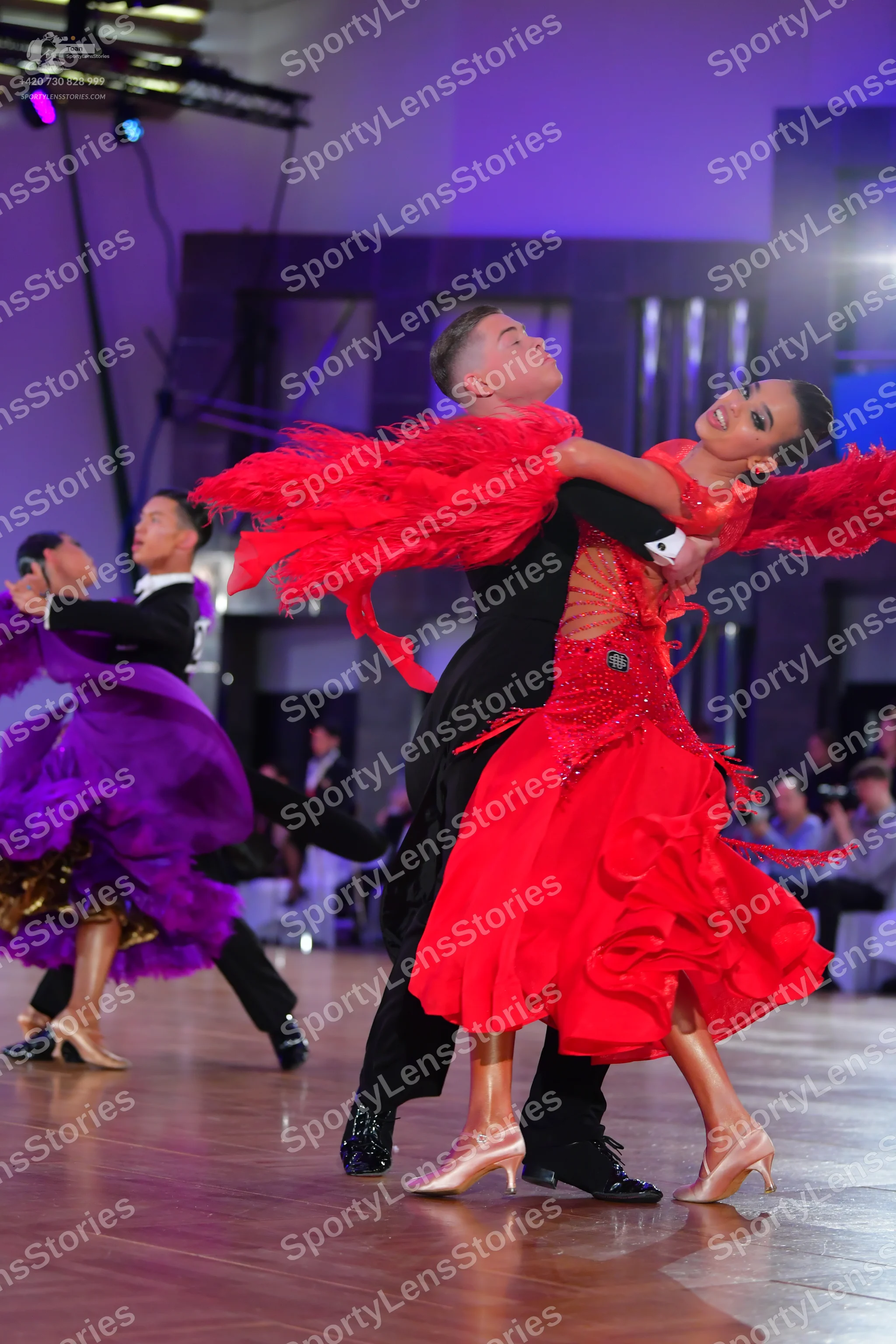 Dancesport competitor: Foreground female dancer: red latin dress with a ruffled skirt and large red fea