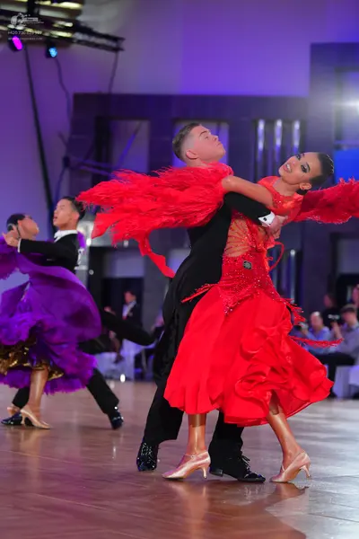 Dancesport competitor: Foreground female dancer: red latin dress with a ruffled skirt and large red fea