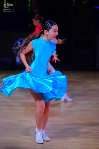 Dancesport competitor: Female dancer in foreground: bright blue sleeveless satin dress with a flared sk