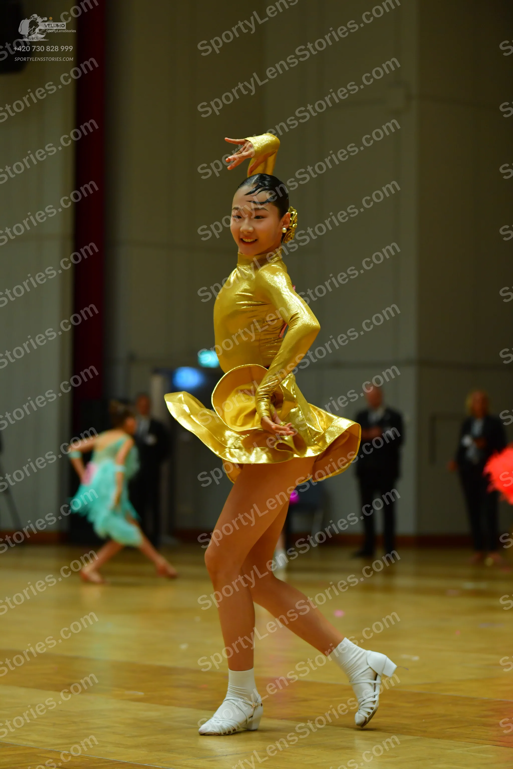 Dancesport competitor: Female dancer in foreground: gold, metallic, long-sleeved Latin dress
