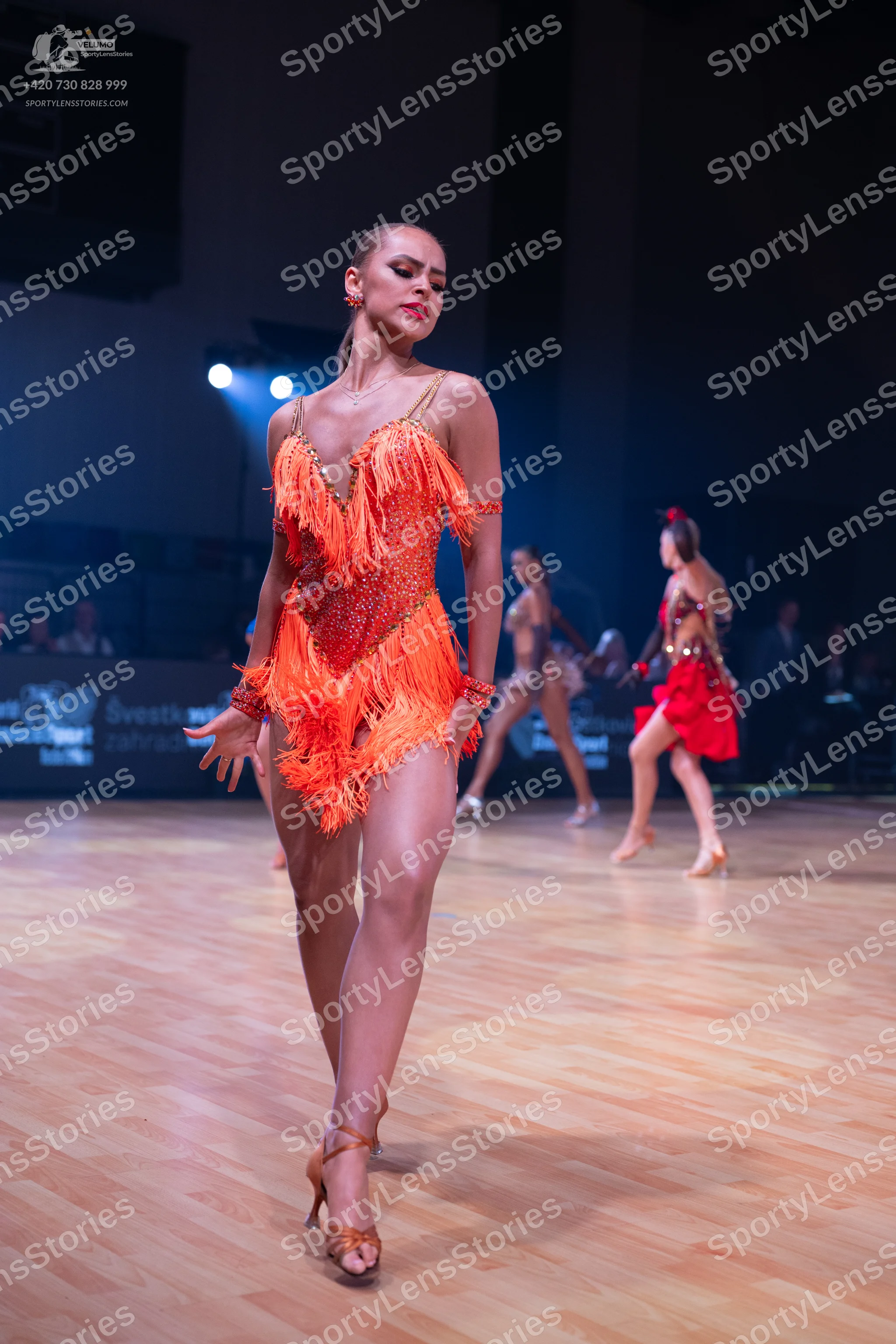 Dancesport competitor: Female dancer in foreground: orange/copper Latin dress with sequins and long fri