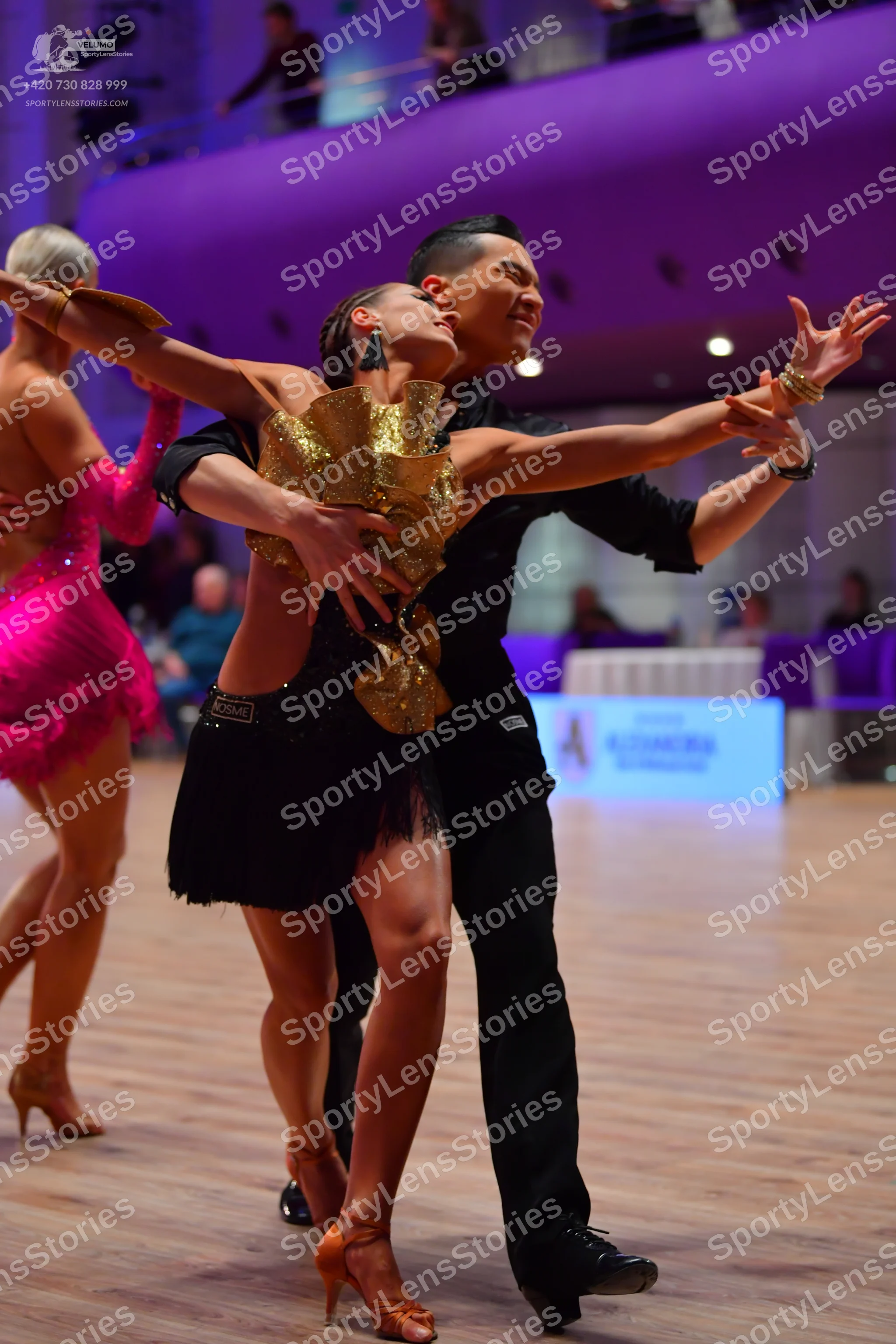 Dancesport competitor: Foreground female dancer: gold sequined and ruffled Latin top with a short black
